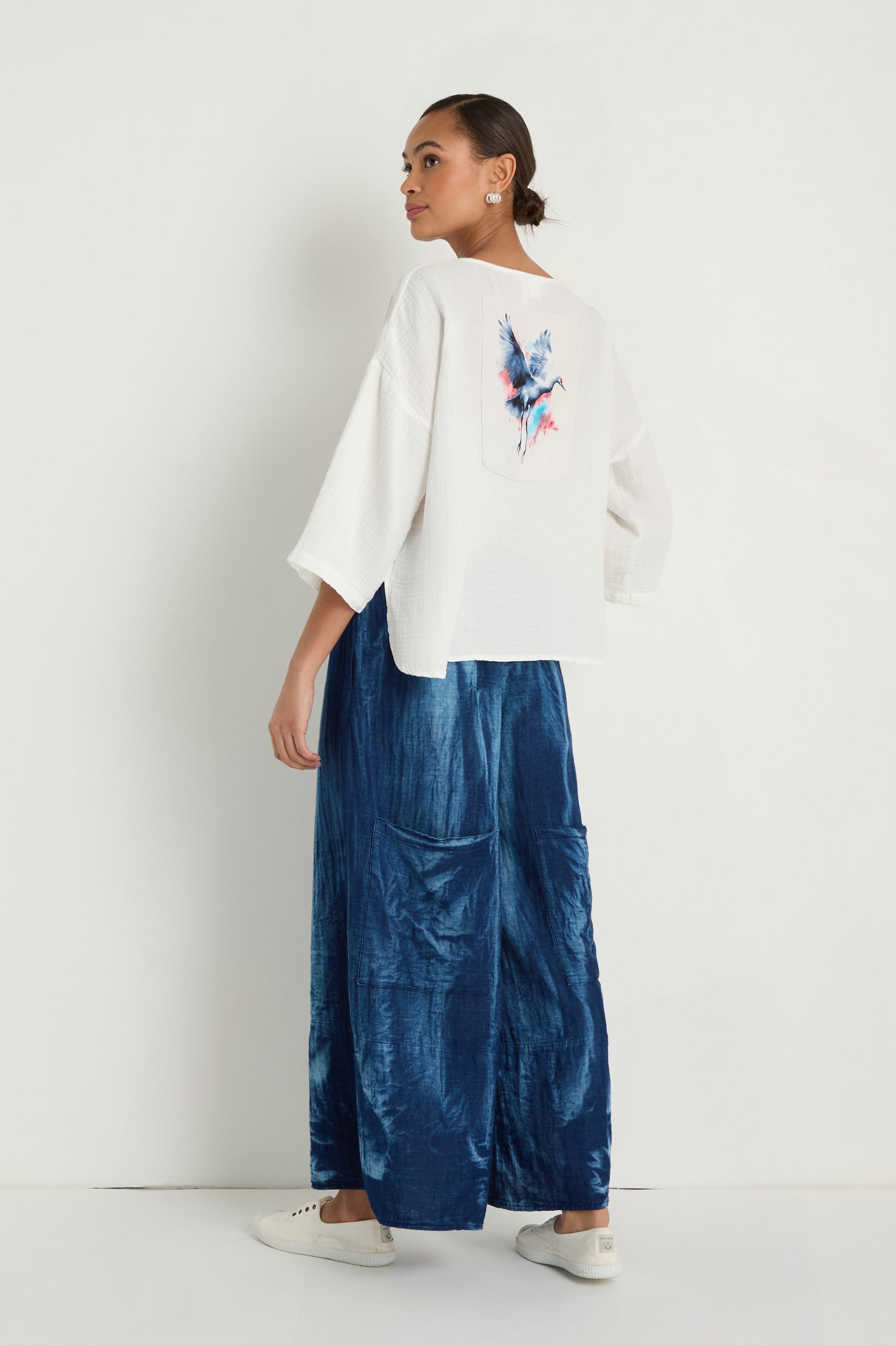 A woman stands sideways in front of a white wall, wearing the Crinkle Two-Pocket Top with a colorful back design, paired with blue wide-leg pants and white sneakers.