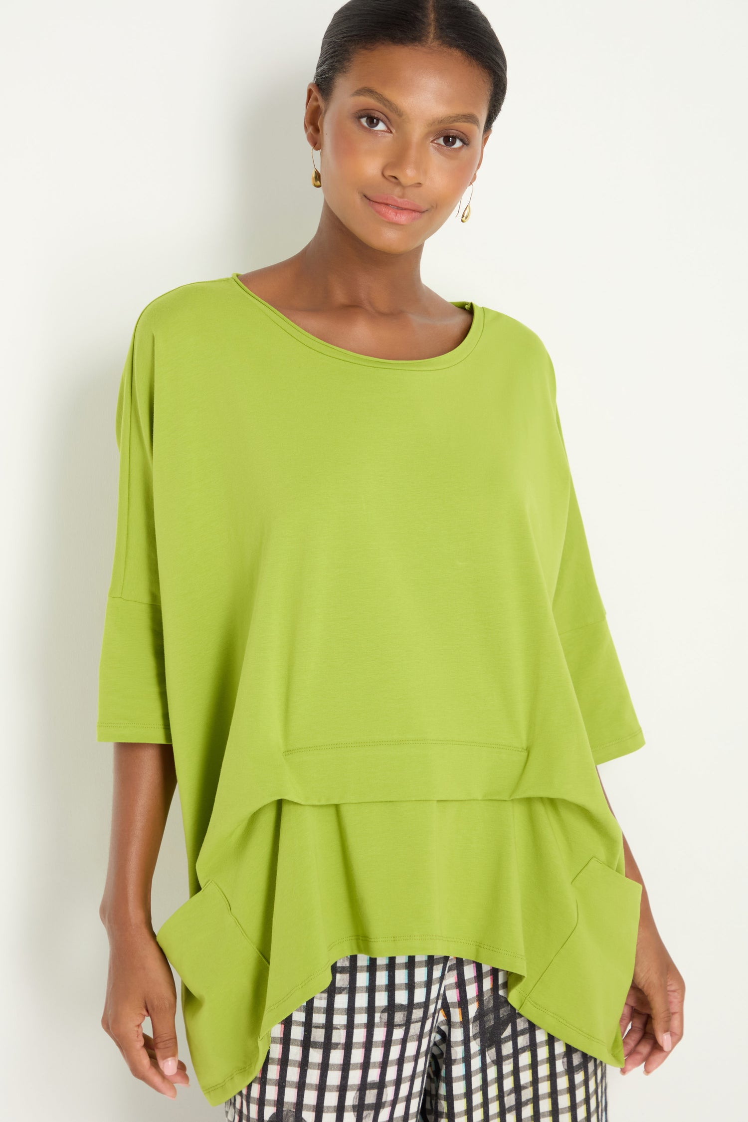 A woman models the Tuck Hem Swing Lime Tunic, a loose-fitting, short-sleeve cotton blend top with two front pockets, paired with black and white checkered pants against a plain white background for easy style and comfort.