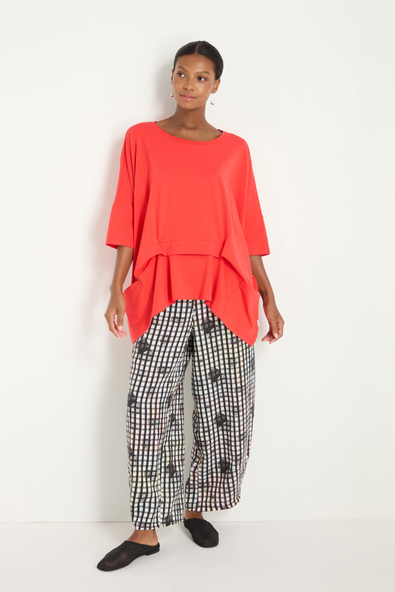 A woman stands against a white wall wearing the Tuck Hem Swing Red Tunic, loose-fitting red cotton blend top, paired with wide-leg black and white grid-patterned pants and black slip-on shoes.