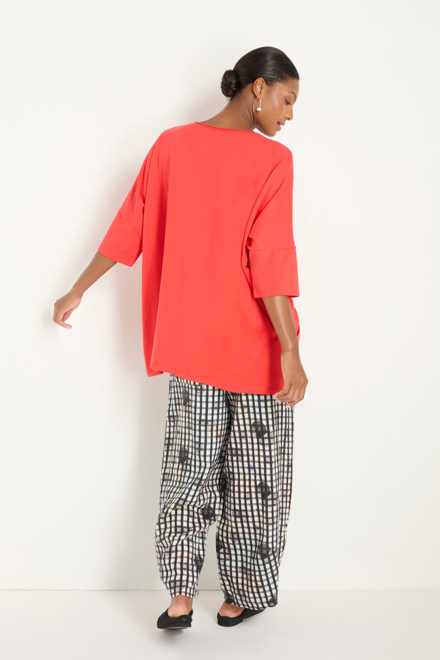 A woman faces away, wearing the Tuck Hem Swing Red Tunic, black and white checkered wide-leg pants with spots, and black flats against a plain white background.