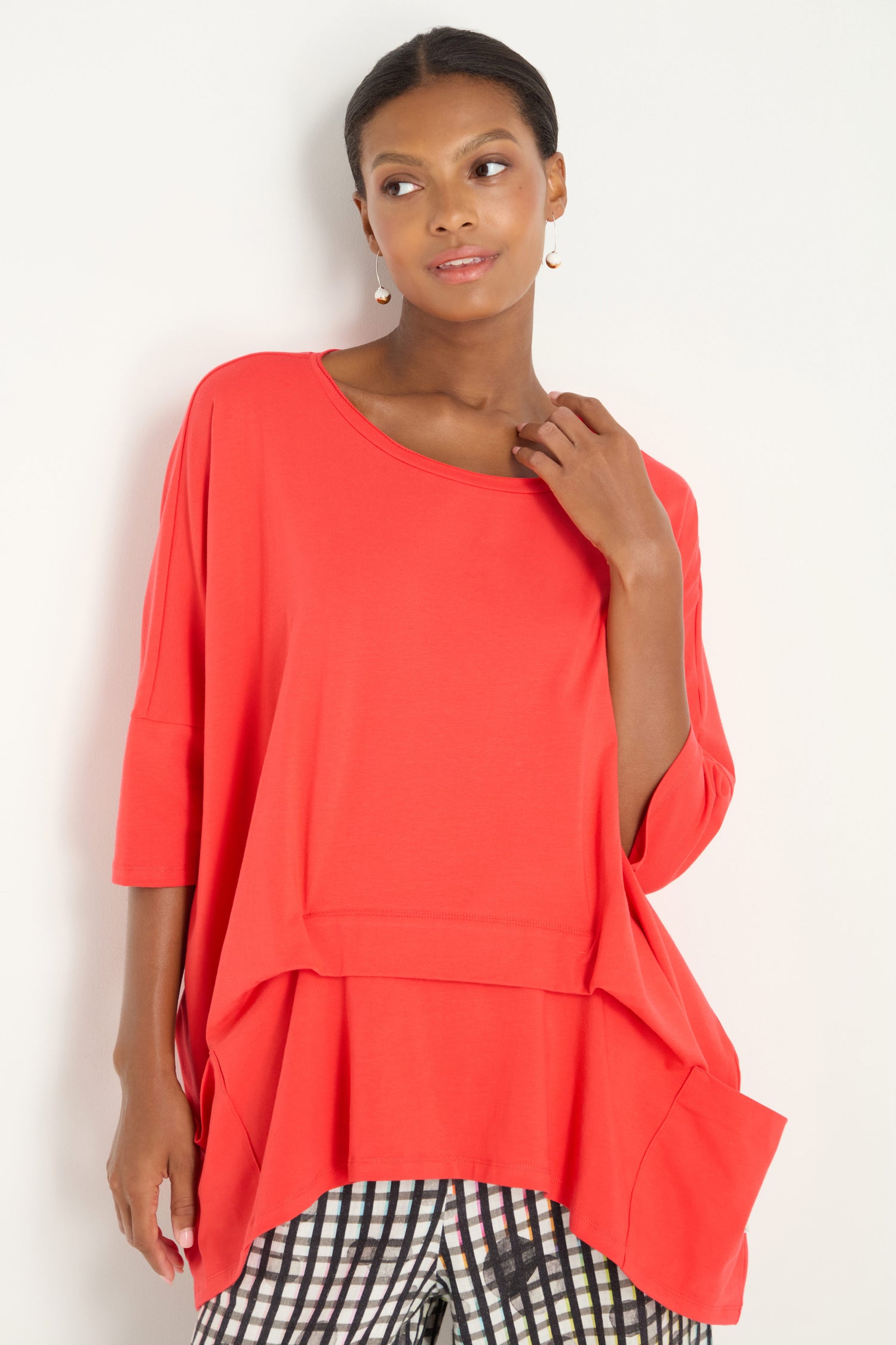 A woman stands against a plain white background wearing the Tuck Hem Swing Red Tunic, a loose-fitting, bright red top with three-quarter sleeves and a tucked hem, paired with black-and-white patterned pants.