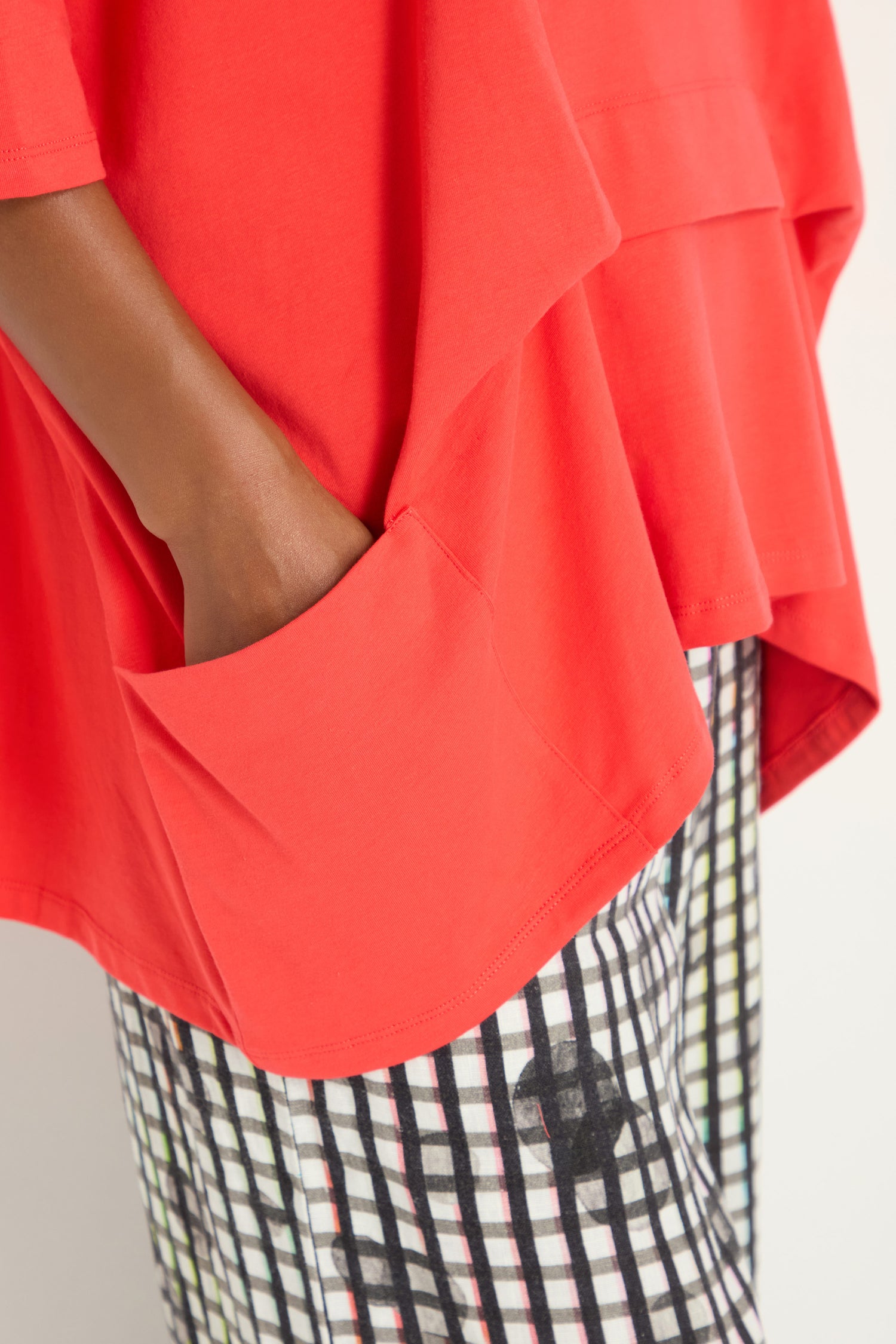 A person wearing a Tuck Hem Swing Red Tunic with a loose fit and large pocket, paired with checkered black-and-white pants, one hand casually in the tunic’s pocket.