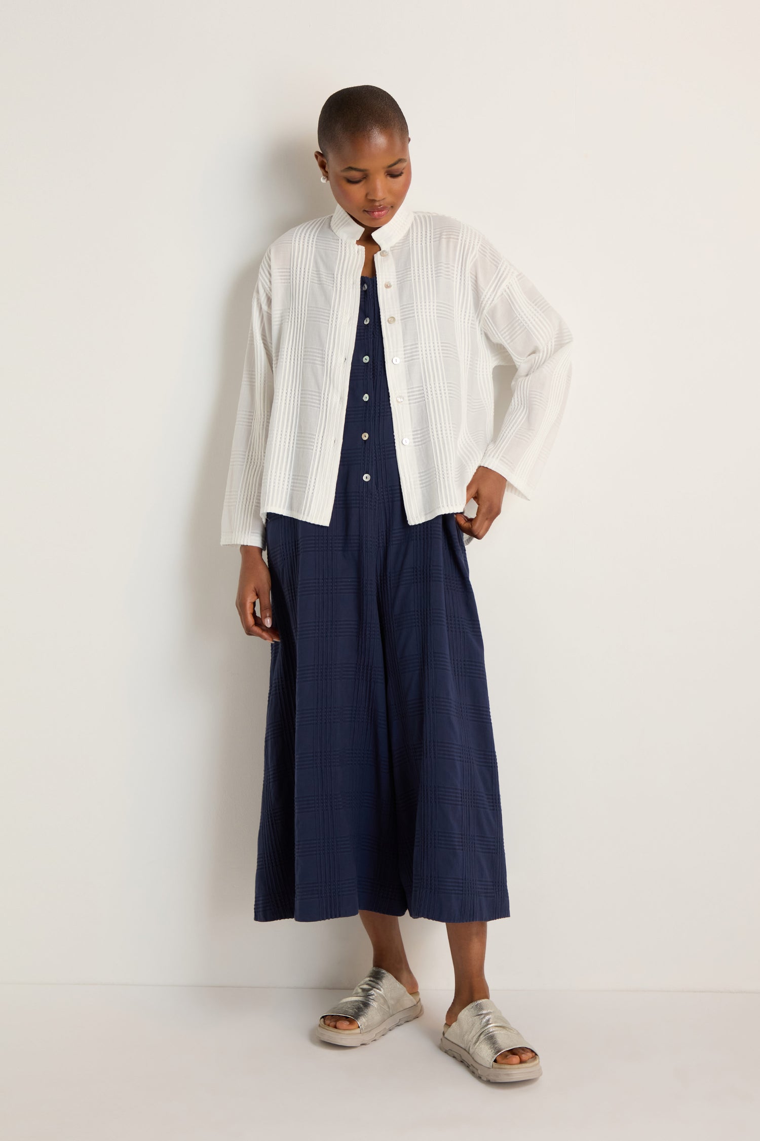 Person stands against a white wall, wearing the Self Check White Boxy Shirt in a relaxed boxy silhouette over a navy blue dress and paired with silver sandals.