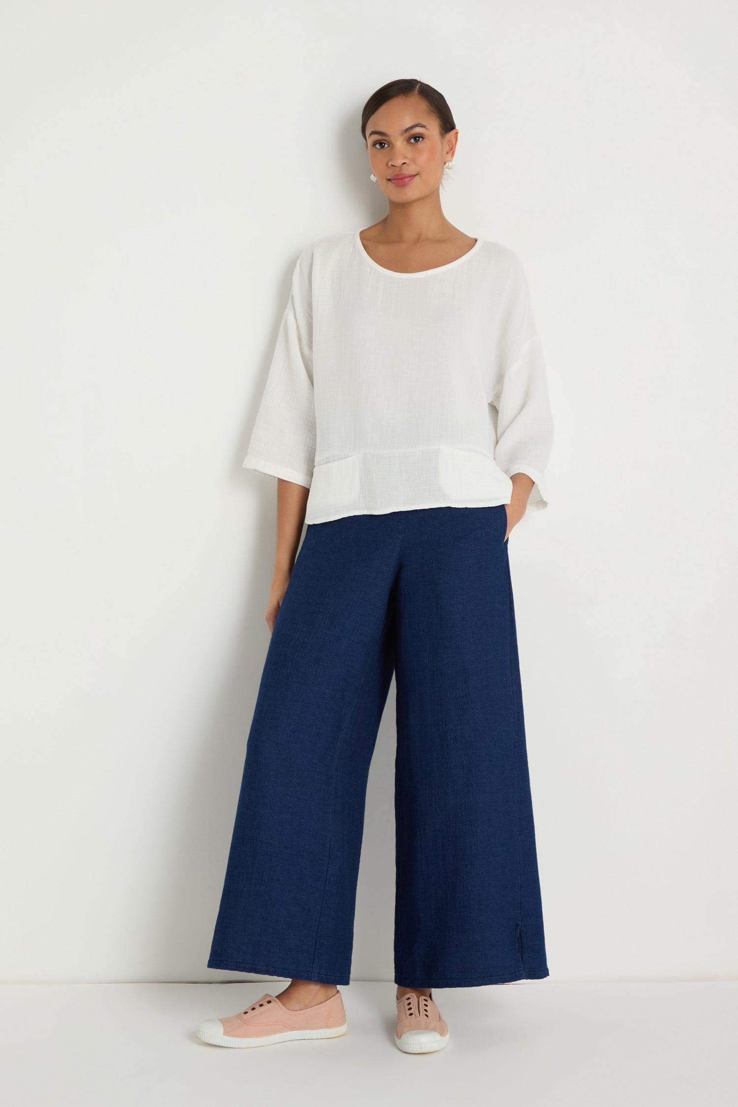 A woman stands against a white wall wearing a loose white top, Wide Leg Slit Trousers in blue, and light pink sneakers.
