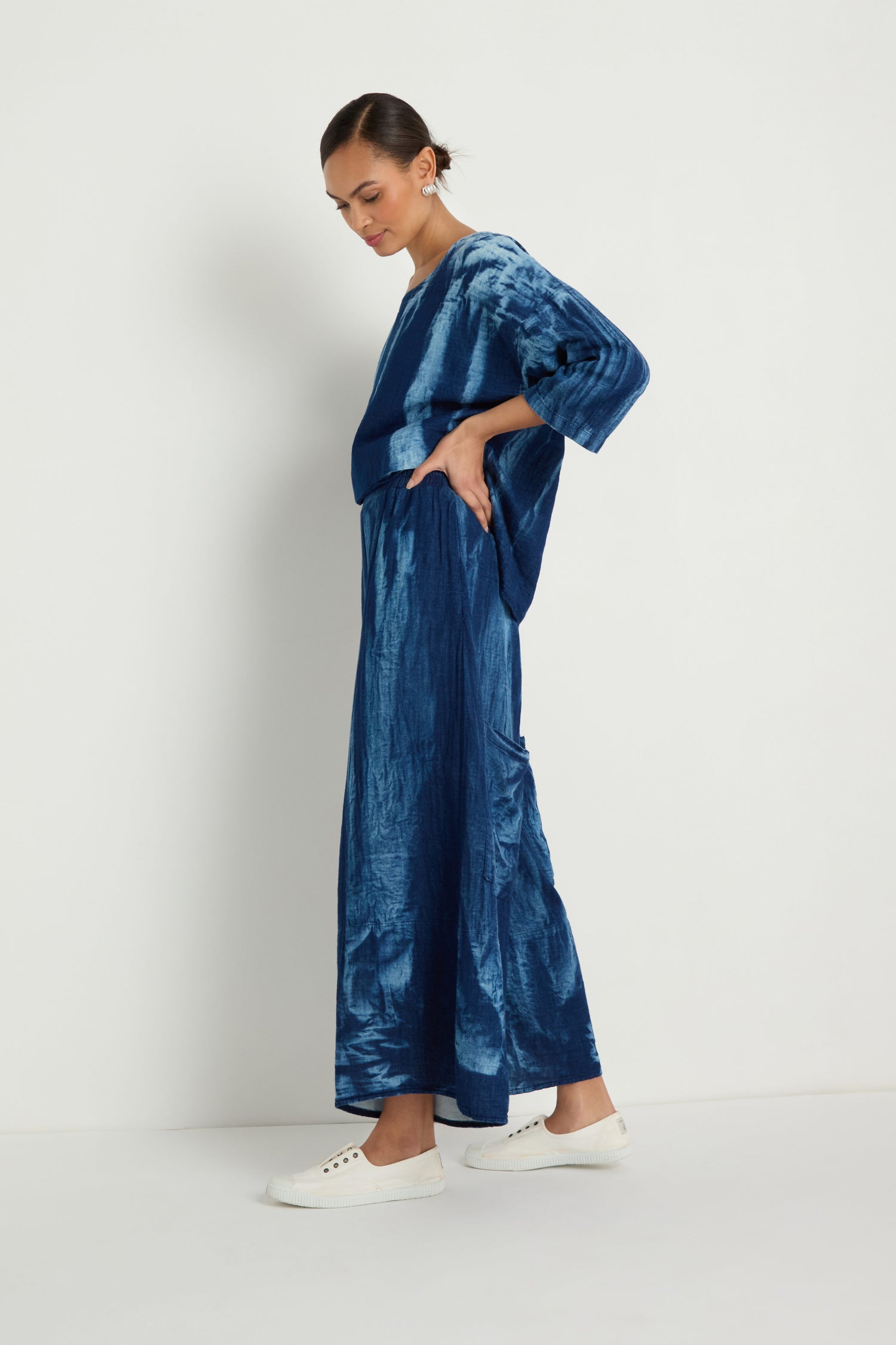 A woman stands sideways against a white background wearing Soft Denim Pocket Trousers in blue tie-dye with a matching loose-fitting top and white sneakers.