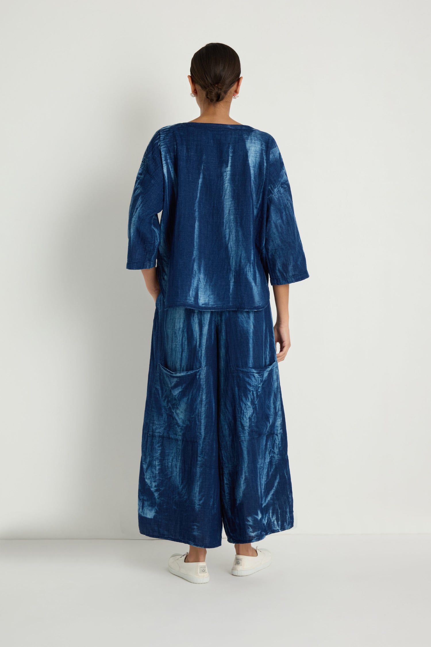 Facing a white wall, a person wears loose blue long sleeves, Soft Denim Pocket Trousers in matching blue, and white shoes.