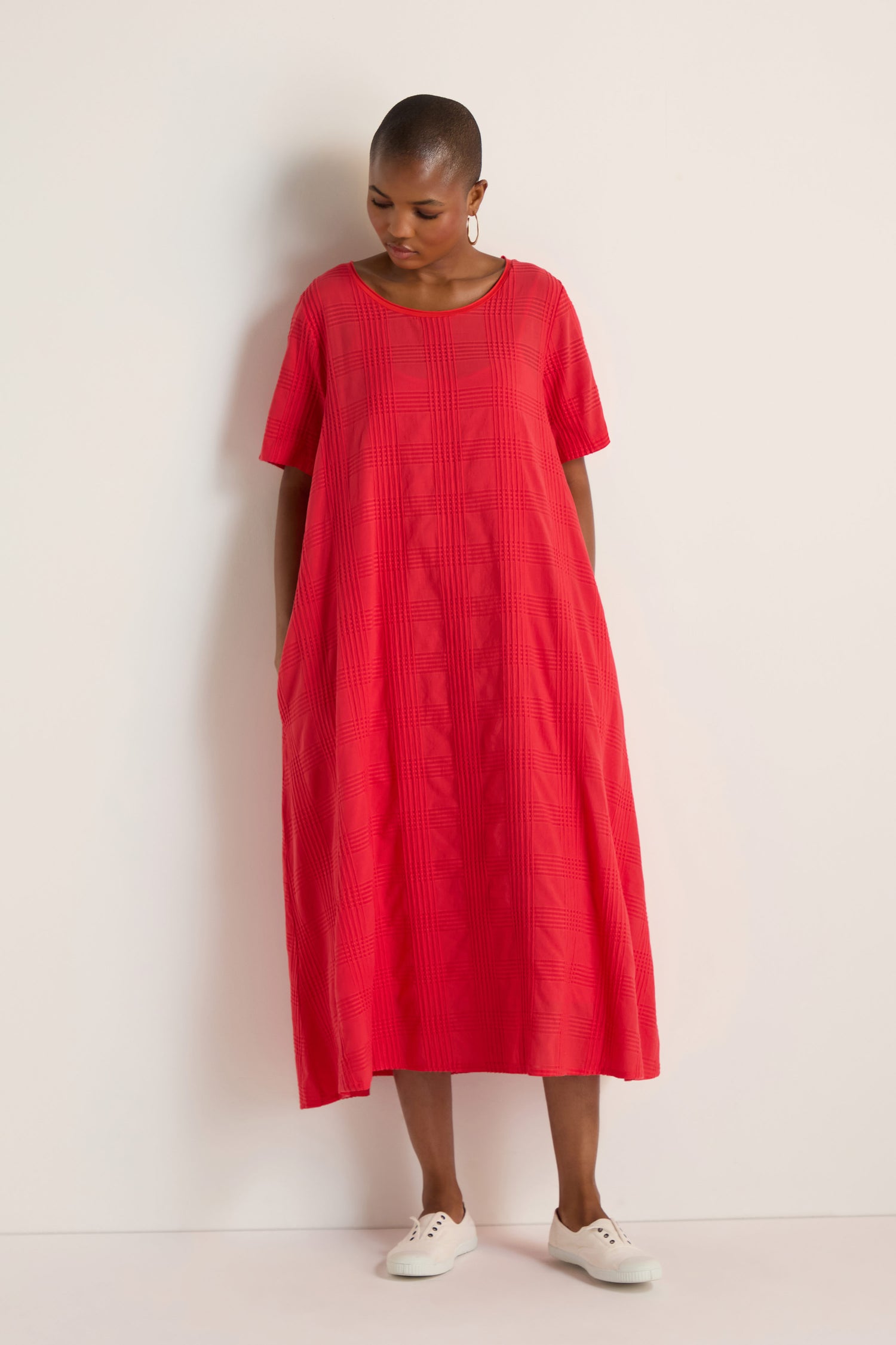 A person stands against a white wall wearing a loose-fitting, short-sleeved Self Check Flared Dress in red and white shoes.