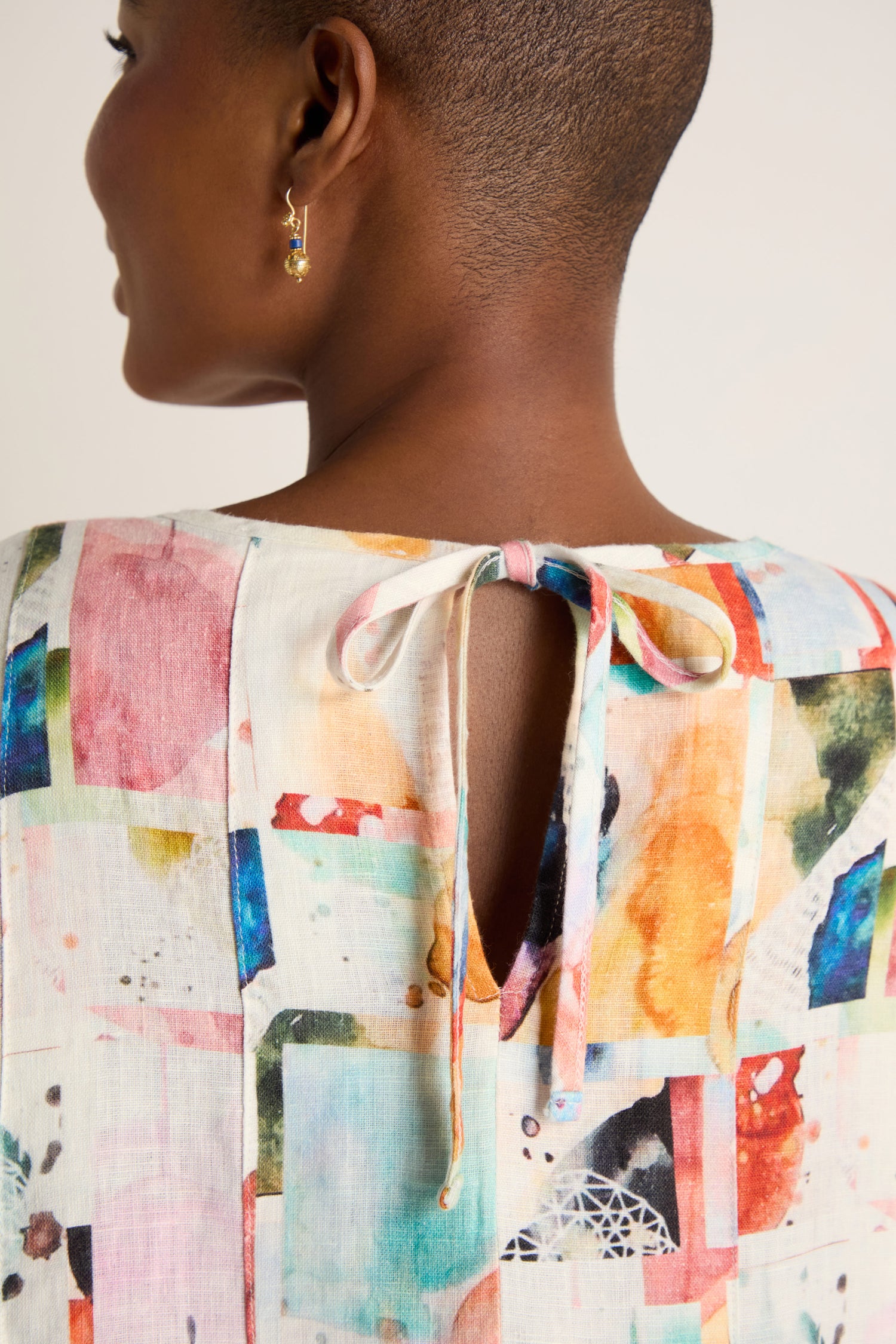 A person with short hair wears the Watercolour Linen Pleated Dress, a lightweight summer style with a colorful patterned top, open back, tie neckline, and visible small hoop earring.