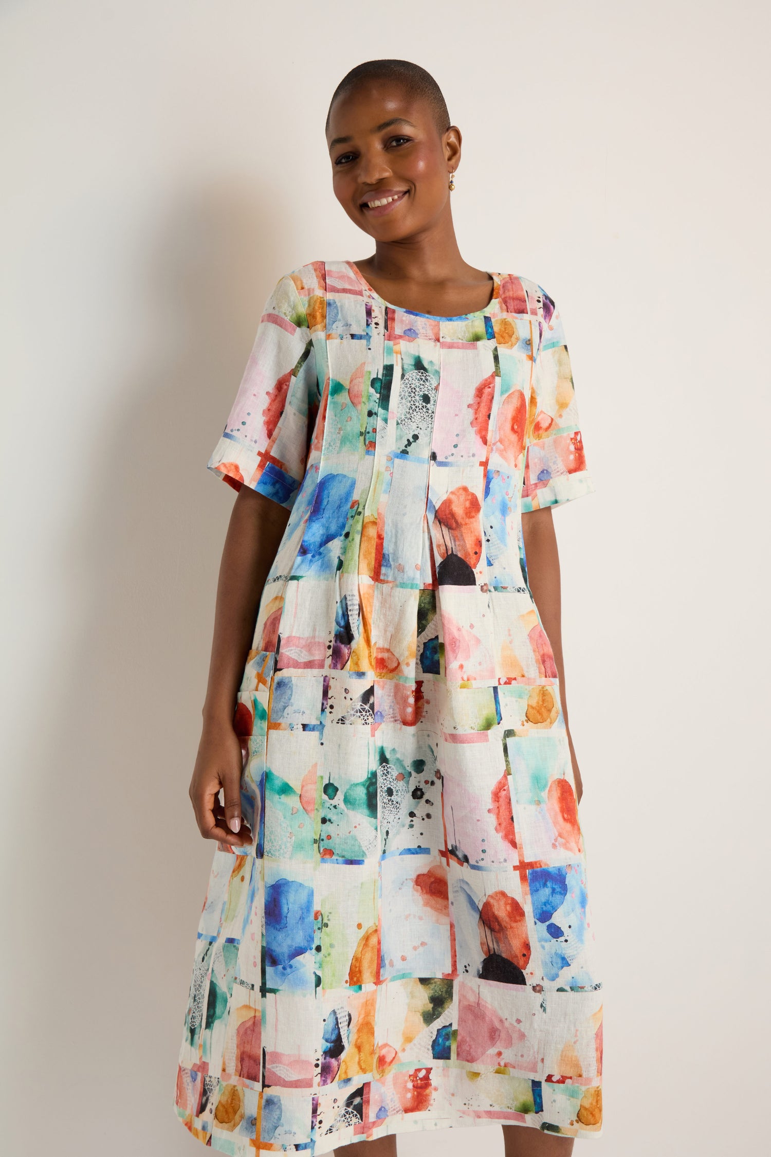 A person with short hair smiles while wearing the Watercolour Linen Pleated Dress, featuring colorful abstract patterns, against a plain light background.