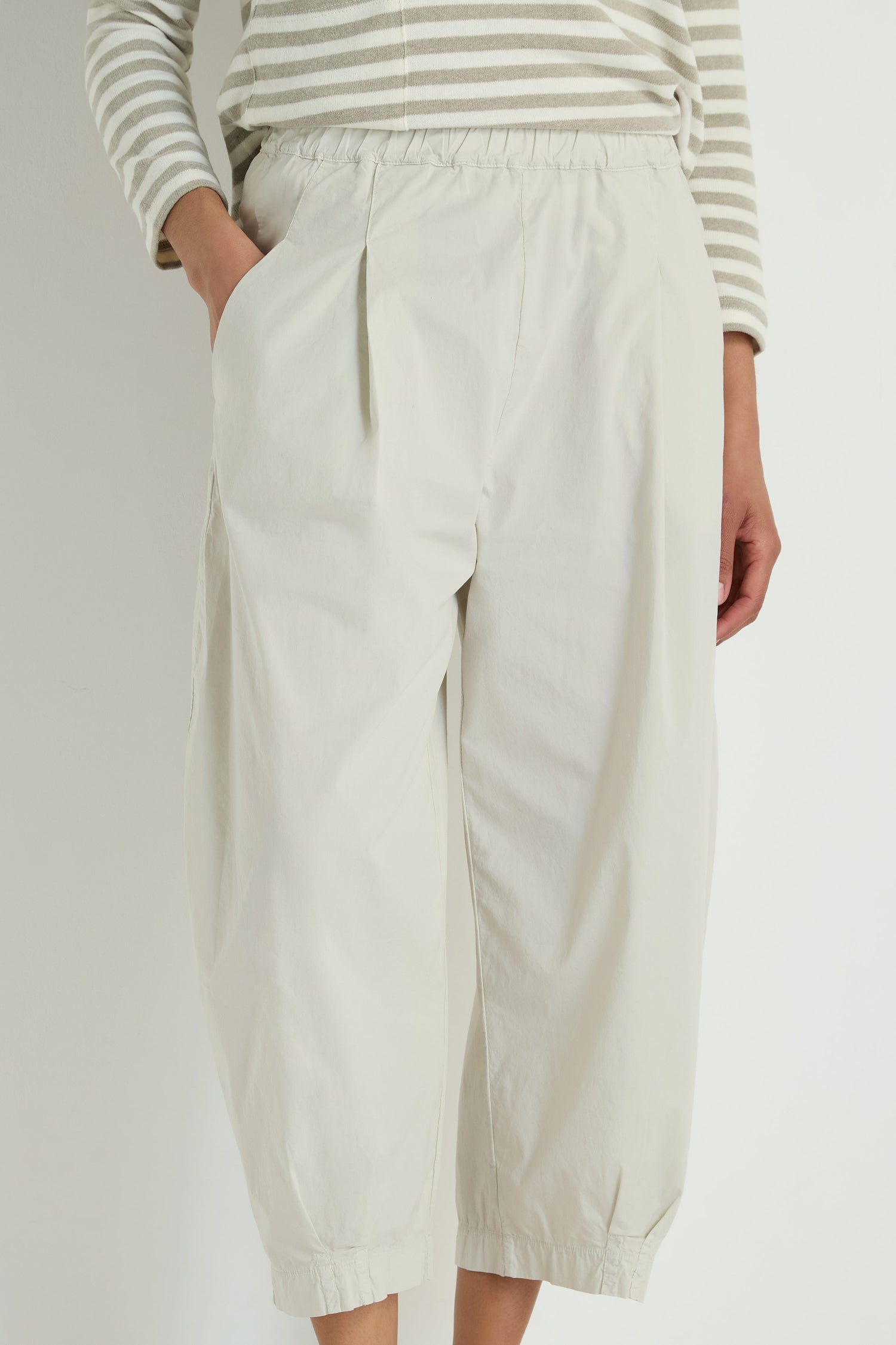 Person wearing Pleated Poplin Trousers in loose, light beige with a tapered leg and hands in pockets, paired with a long-sleeve light gray and white striped cotton blend poplin top.