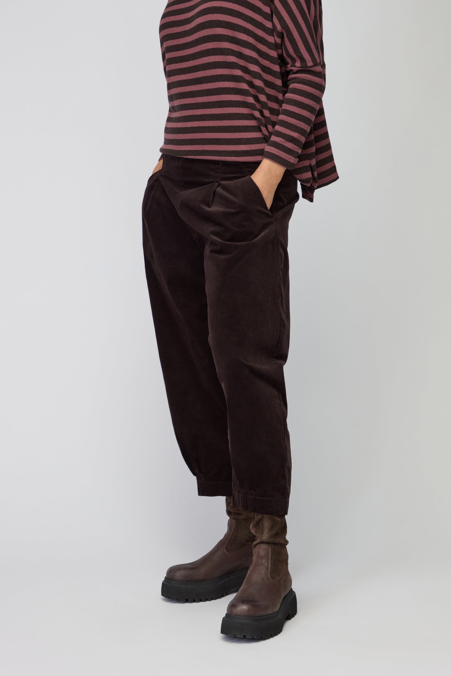 Person wearing Fine Cord Stretch Trousers in dark brown with an elasticated waistband, paired with a matching long-sleeve top and black ankle boots, standing against a plain background.
