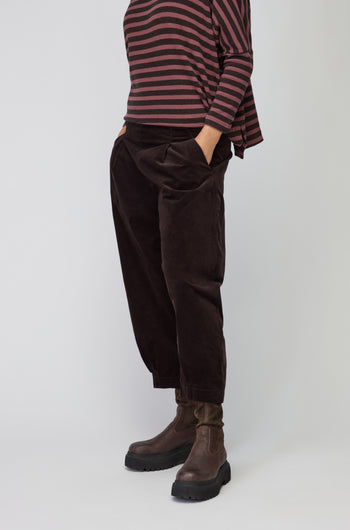 Person wearing Fine Cord Stretch Trousers in dark brown with an elasticated waistband, paired with a matching long-sleeve top and black ankle boots, standing against a plain background.