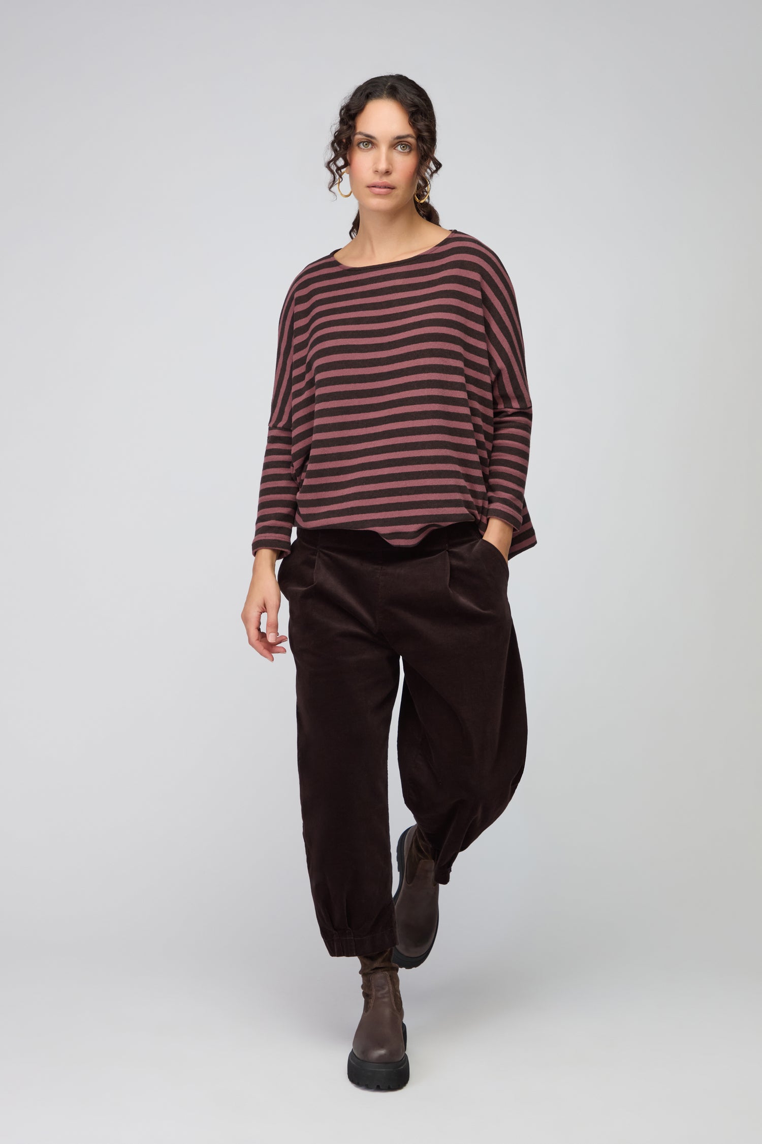 A woman stands against a plain background wearing a loose brown sweater, Fine Cord Stretch Trousers with an elasticated waistband, and black ankle boots, her hands in her pockets and head slightly tilted down.