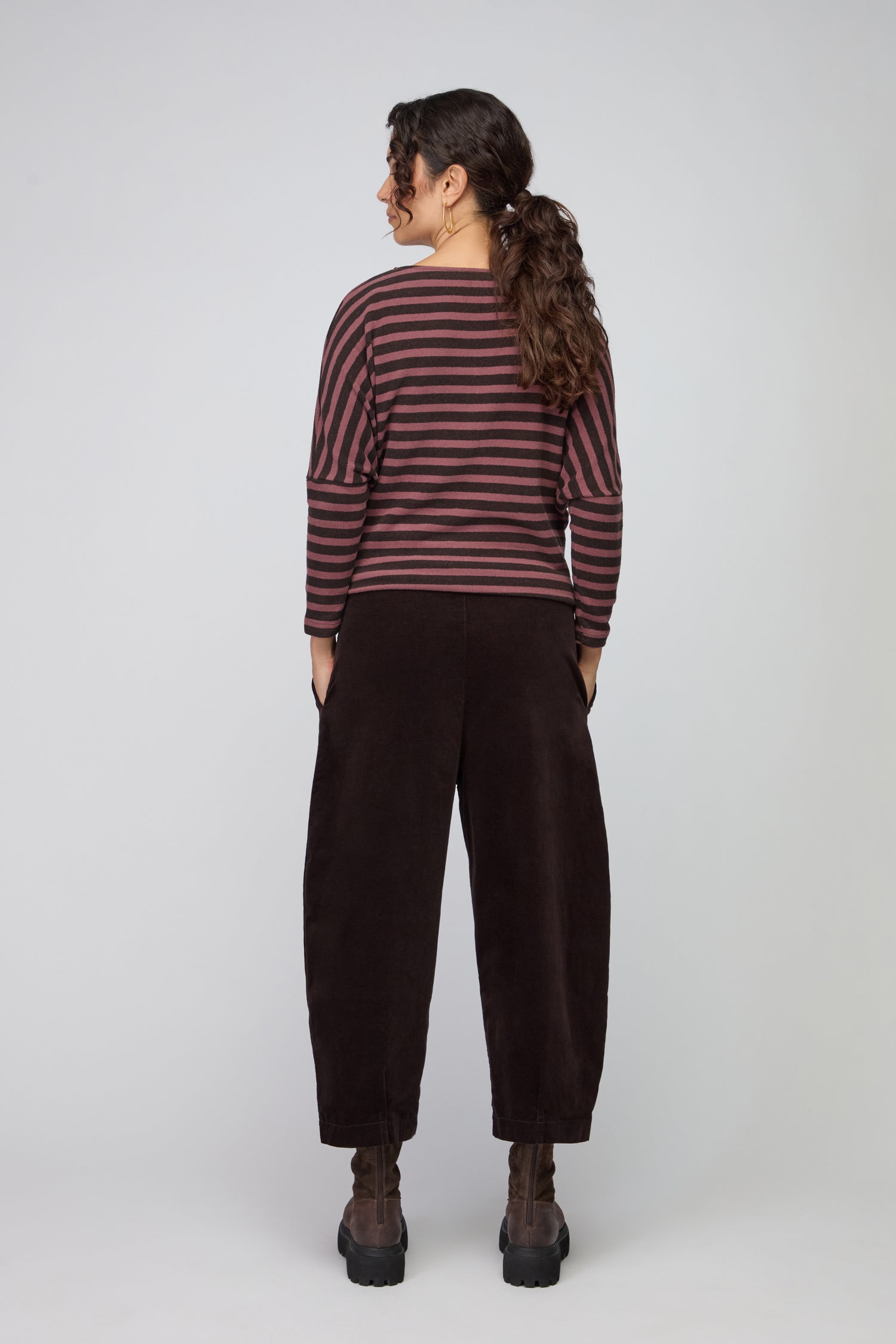 A person stands facing away from the camera, wearing a dark brown long-sleeve top, Fine Cord Stretch Trouser with an elasticated waistband, black boots, and large hoop earrings.