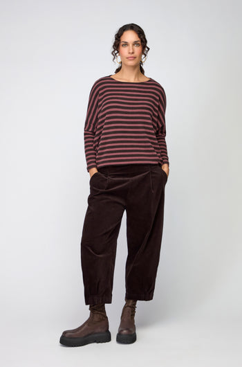 A woman stands against a plain background wearing loose brown sweater, Fine Cord Stretch Trouser with an elasticated waistband, black ankle boots, and her hands in her pockets.