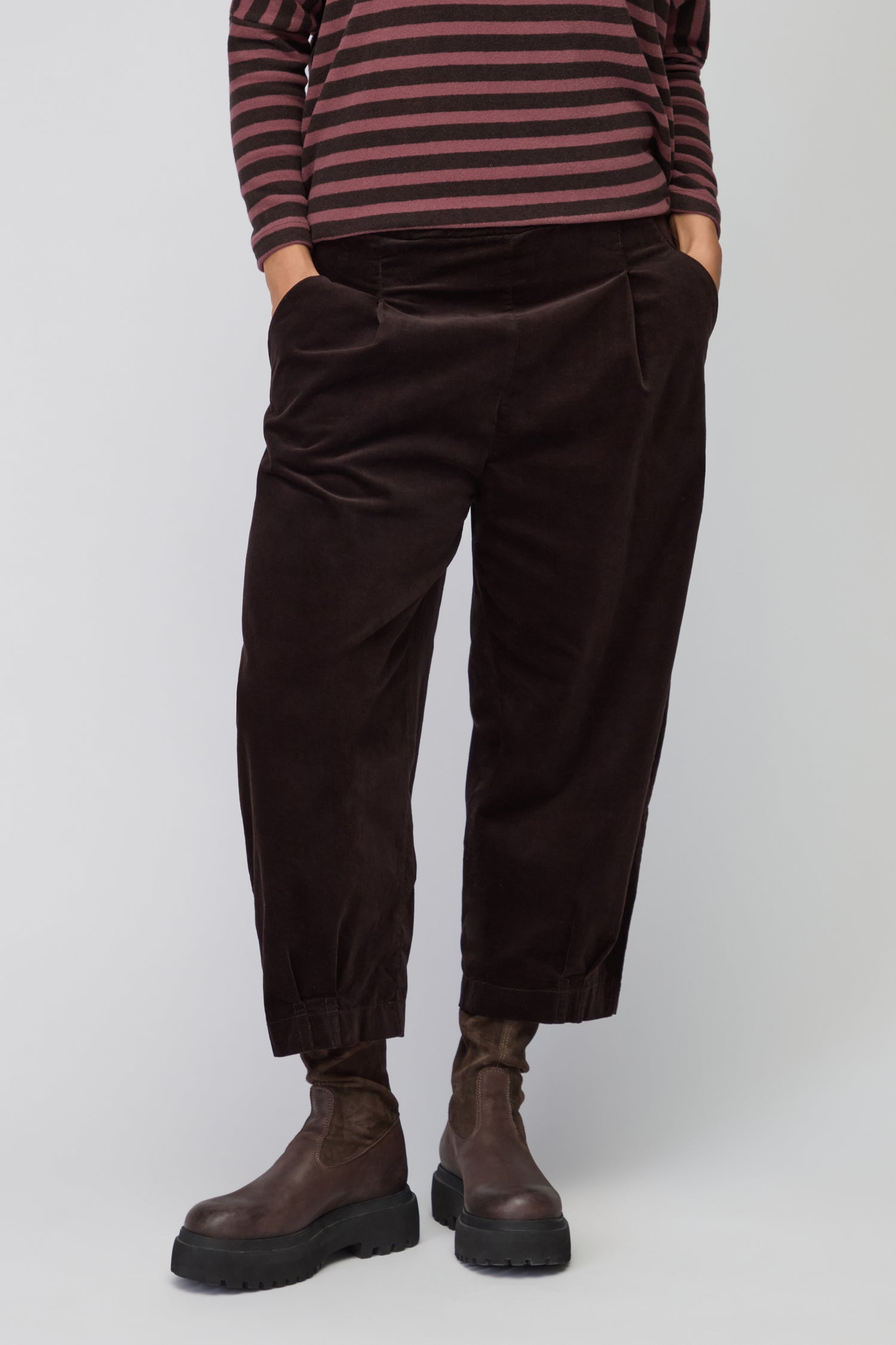 Model wears Fine Cord Stretch Trouser in dark brown with an elasticated waistband, paired with a dark long-sleeve top and black ankle boots, standing against a plain light background.