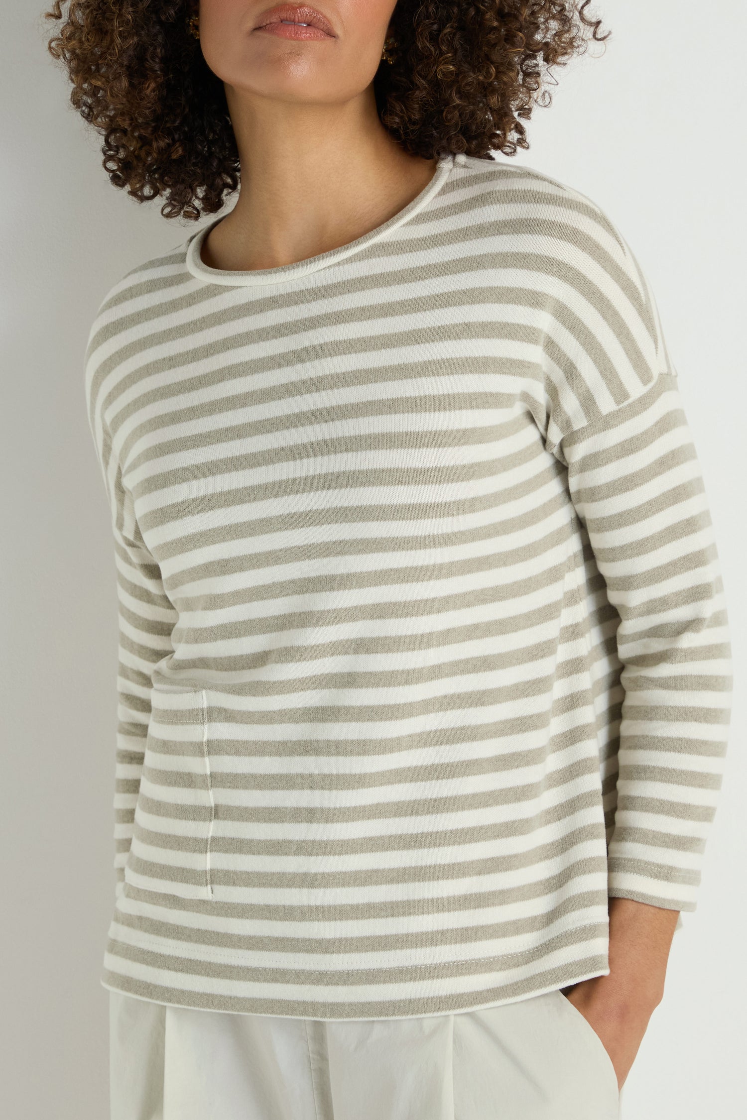 Someone wearing the Stripe Pocket Fleece Top in light beige and white stripes with a relaxed fit, paired with white pants, stands against a plain background.
