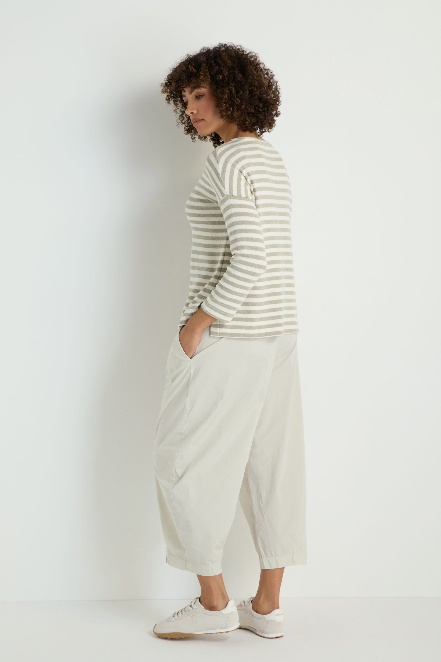 A person stands sideways against a plain background, wearing the Stripe Pocket Fleece Top in beige and white stripes, paired with loose light-colored pants and white sneakers.
