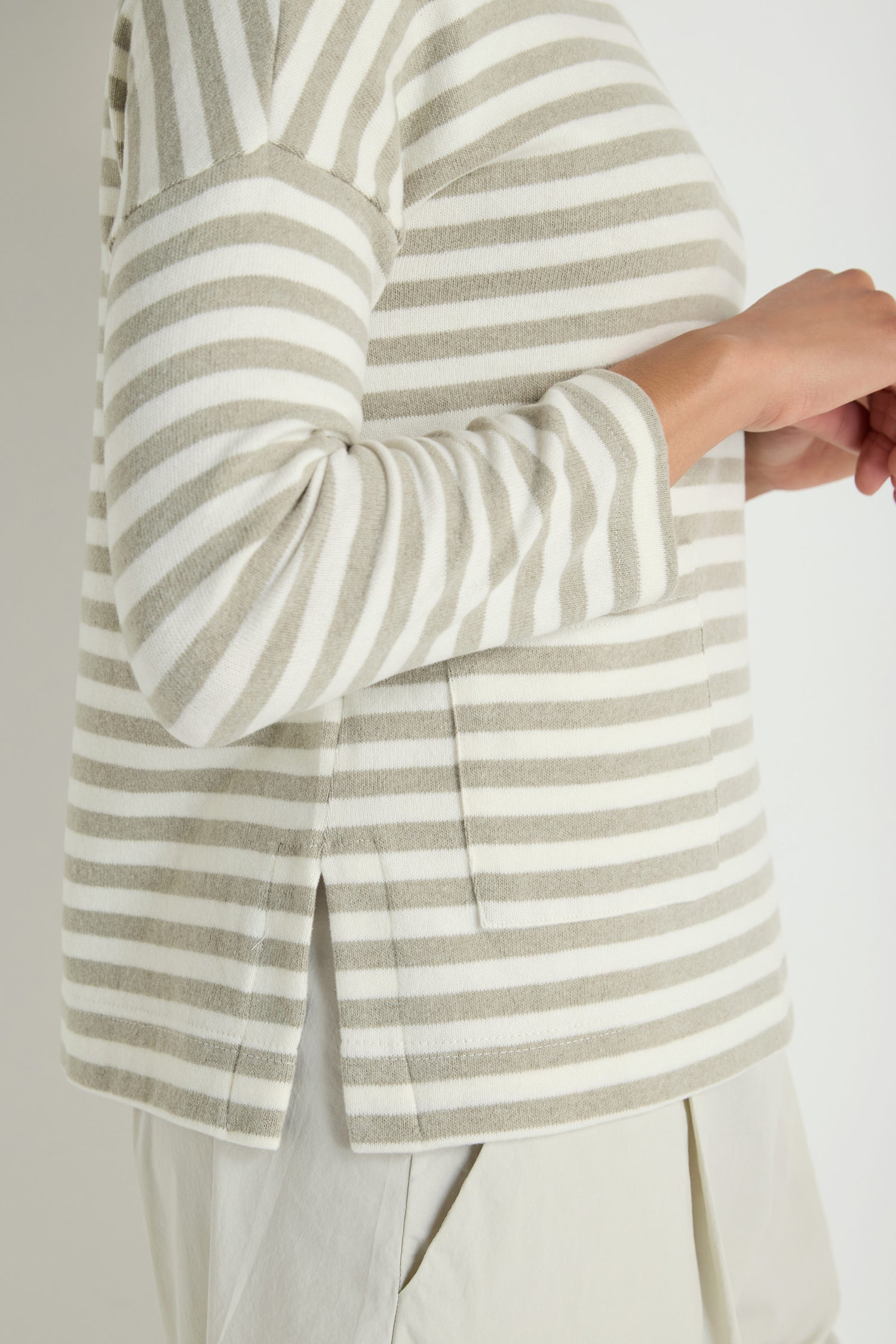 A person wears the Stripe Pocket Fleece Top, featuring a relaxed beige and white striped design, paired with light pants and standing with hands clasped.