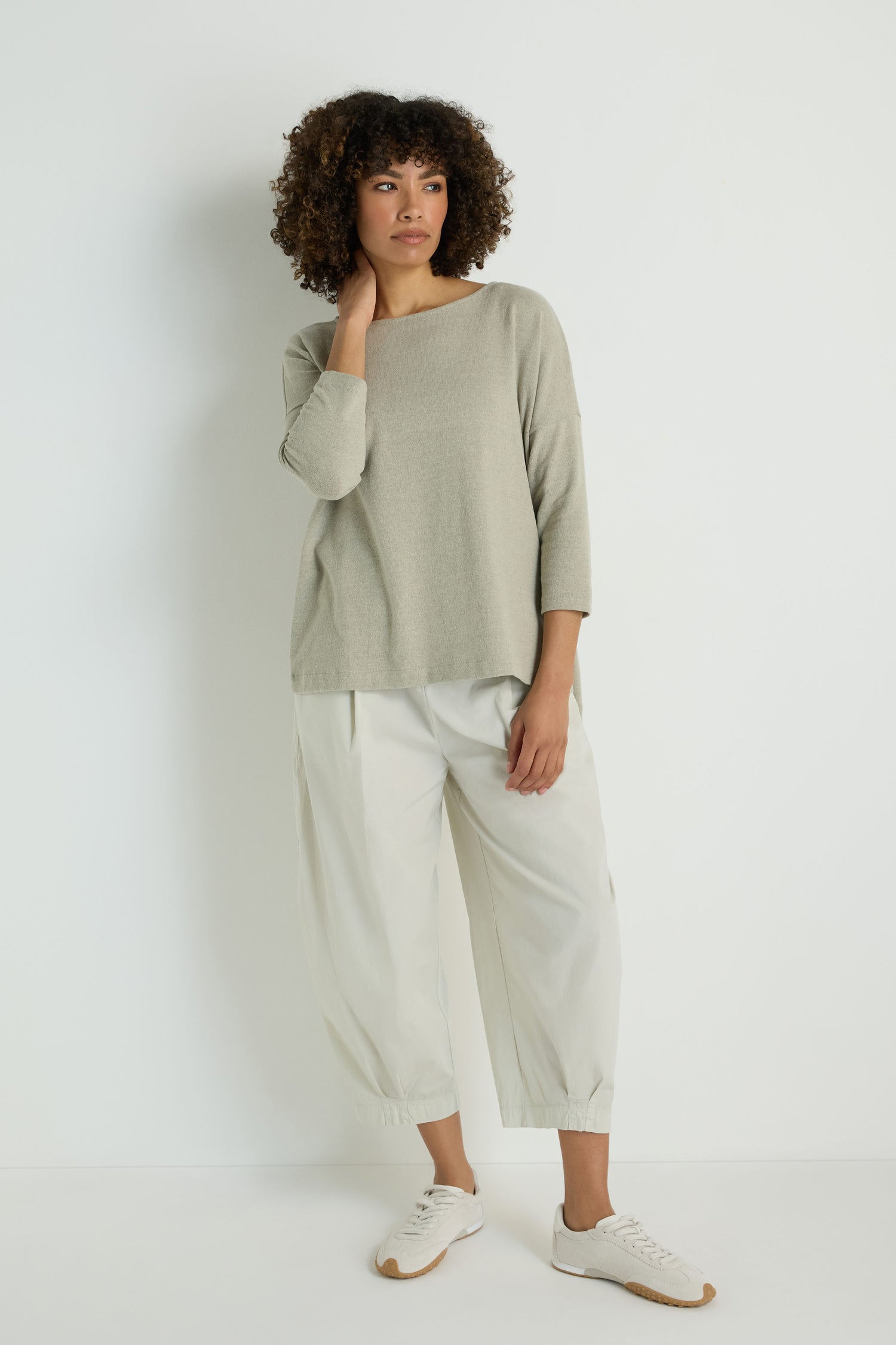A woman stands against a white wall wearing the Flared Fleece Top in light gray, paired with loose white pants and white sneakers.