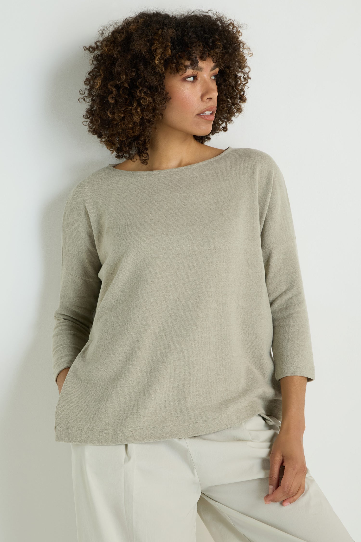 A woman with curly hair models the Flared Fleece Top in light beige, paired with light pants, standing against a plain white background.