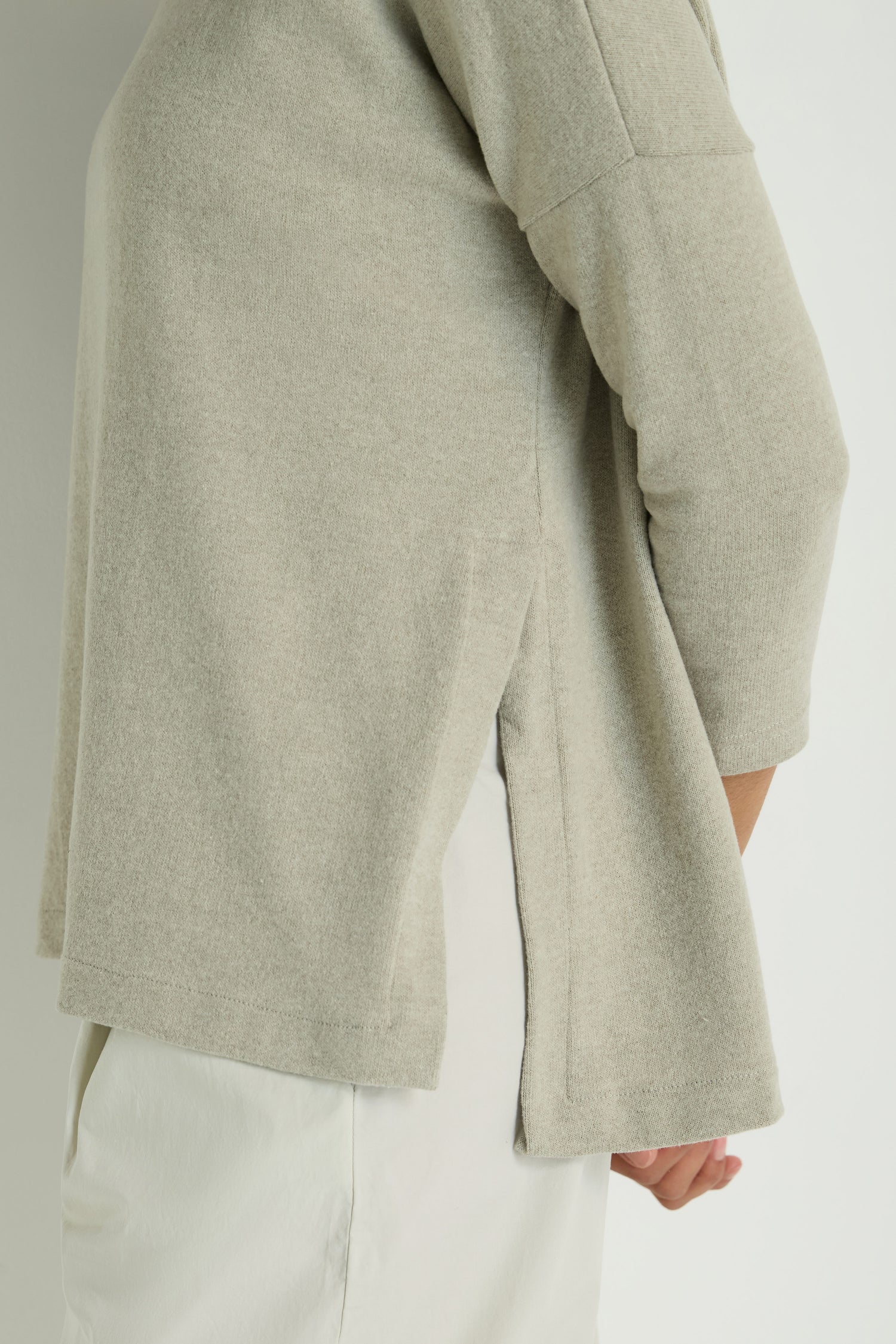 Person wearing the Flared Fleece Top, a light beige cotton-blend, long-sleeve sweater with side split hem, paired with light-colored pants against a plain background.