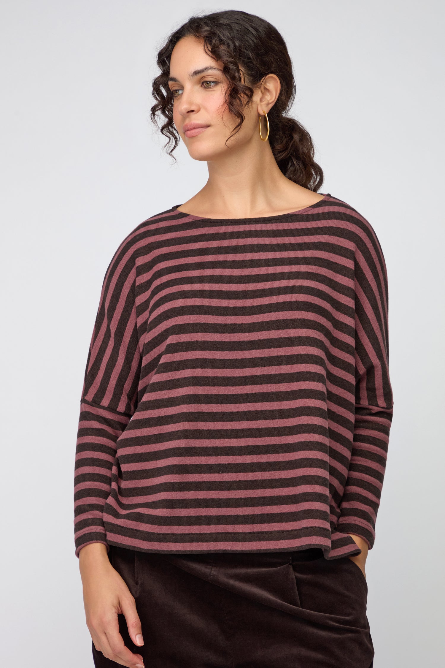 A woman stands slightly sideways, wearing a loose Martis Scoop Neck Wide Stripe Top with wide-leg dark trousers and black boots, set against a plain light background.