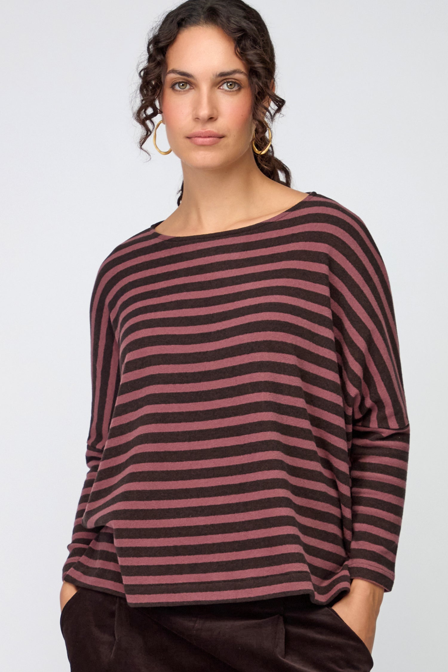 Person wearing a long-sleeve, brown and pink Martis Scoop Neck Wide Stripe Top with side slit and scoop neck, paired with dark gray textured pants, arms crossed.