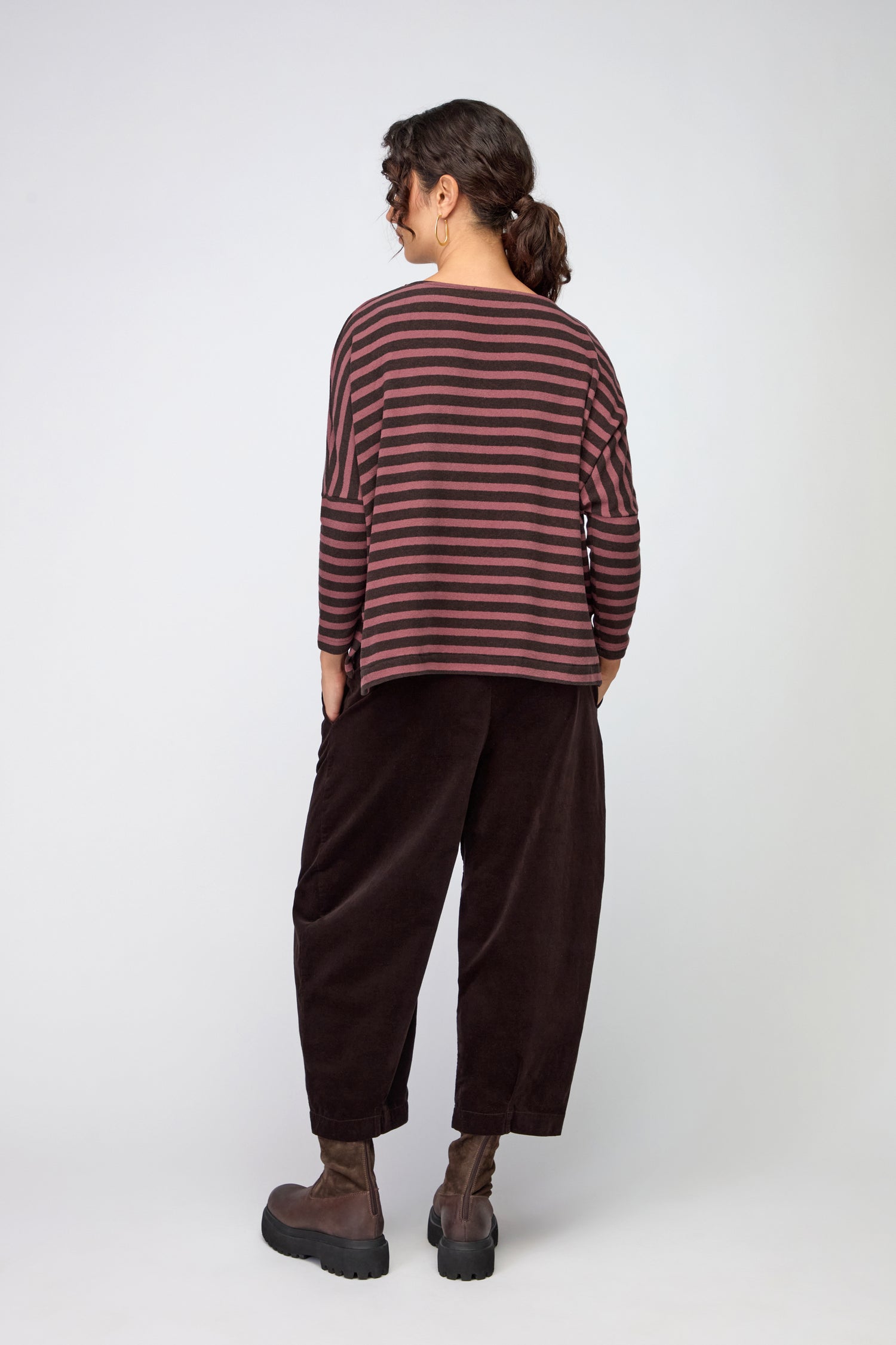 A person stands facing away, wearing a Martis Scoop Neck Wide Stripe Top with red and black stripes, loose dark pants, black boots, and hair tied in a ponytail.