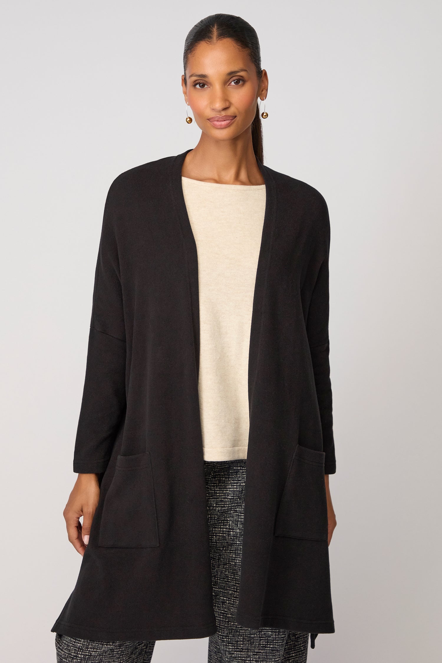 A woman stands facing forward against a plain light background, wearing the black Souffle Soft Jersey Cardi layered over a beige top and patterned pants.