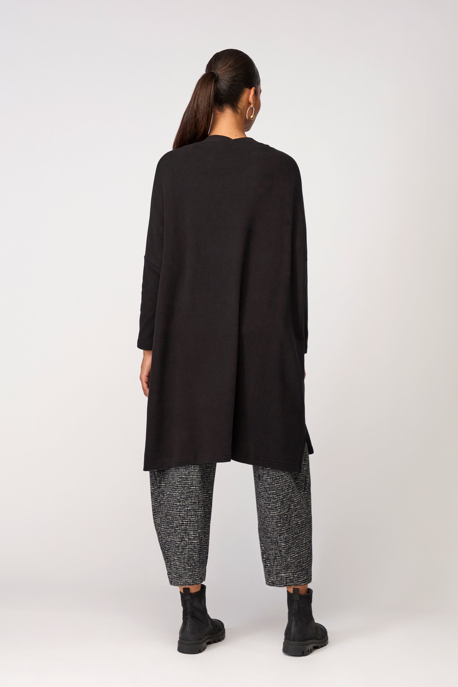 A person stands facing away against a plain background, wearing the black Souffle Soft Jersey Cardi from Mama B over gray textured pants, paired with black boots and their hair in a ponytail.