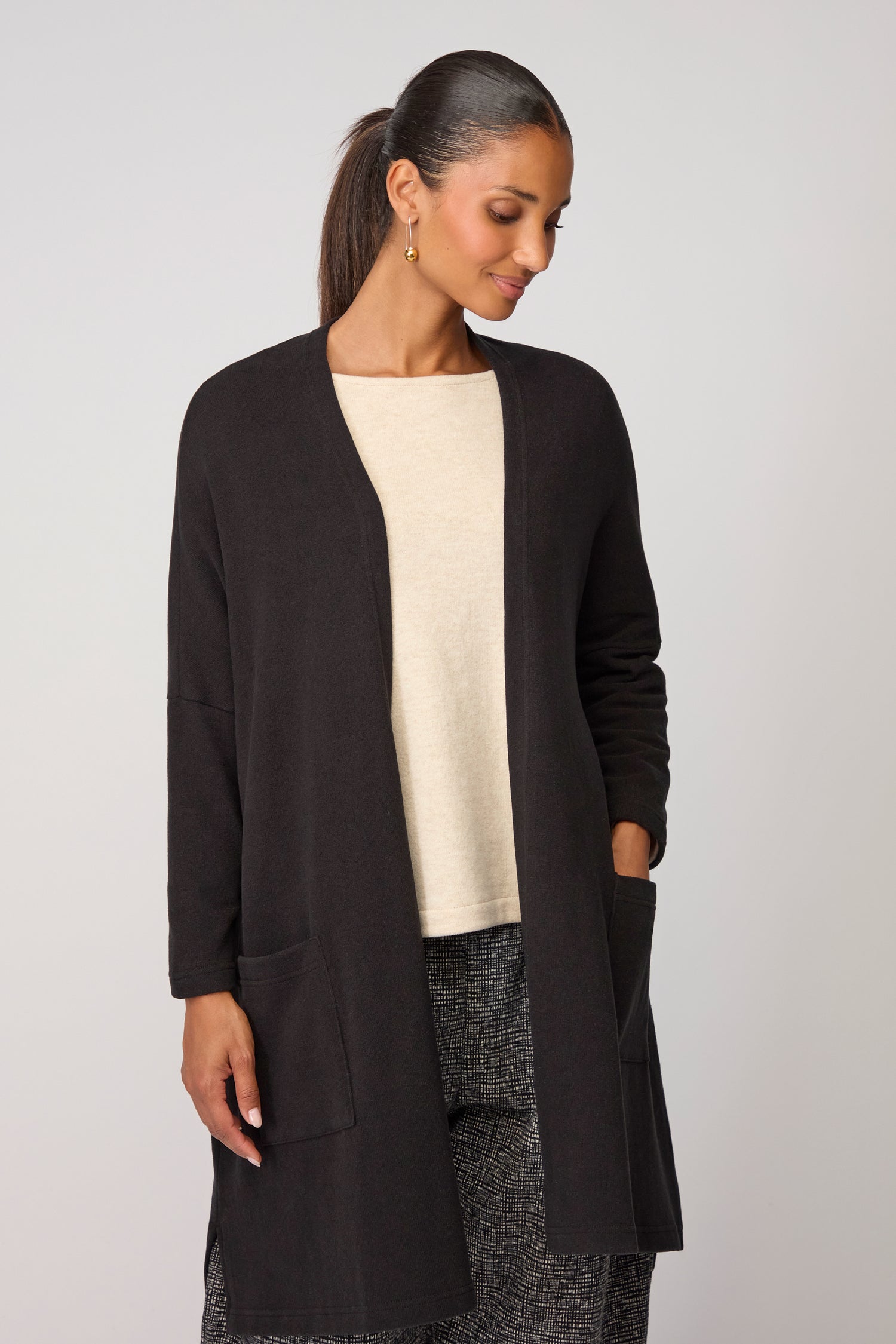 A woman with dark hair in a ponytail wears the black Souffle Soft Jersey Cardi—a versatile layering piece—over a beige top and patterned pants, standing against a plain light background.