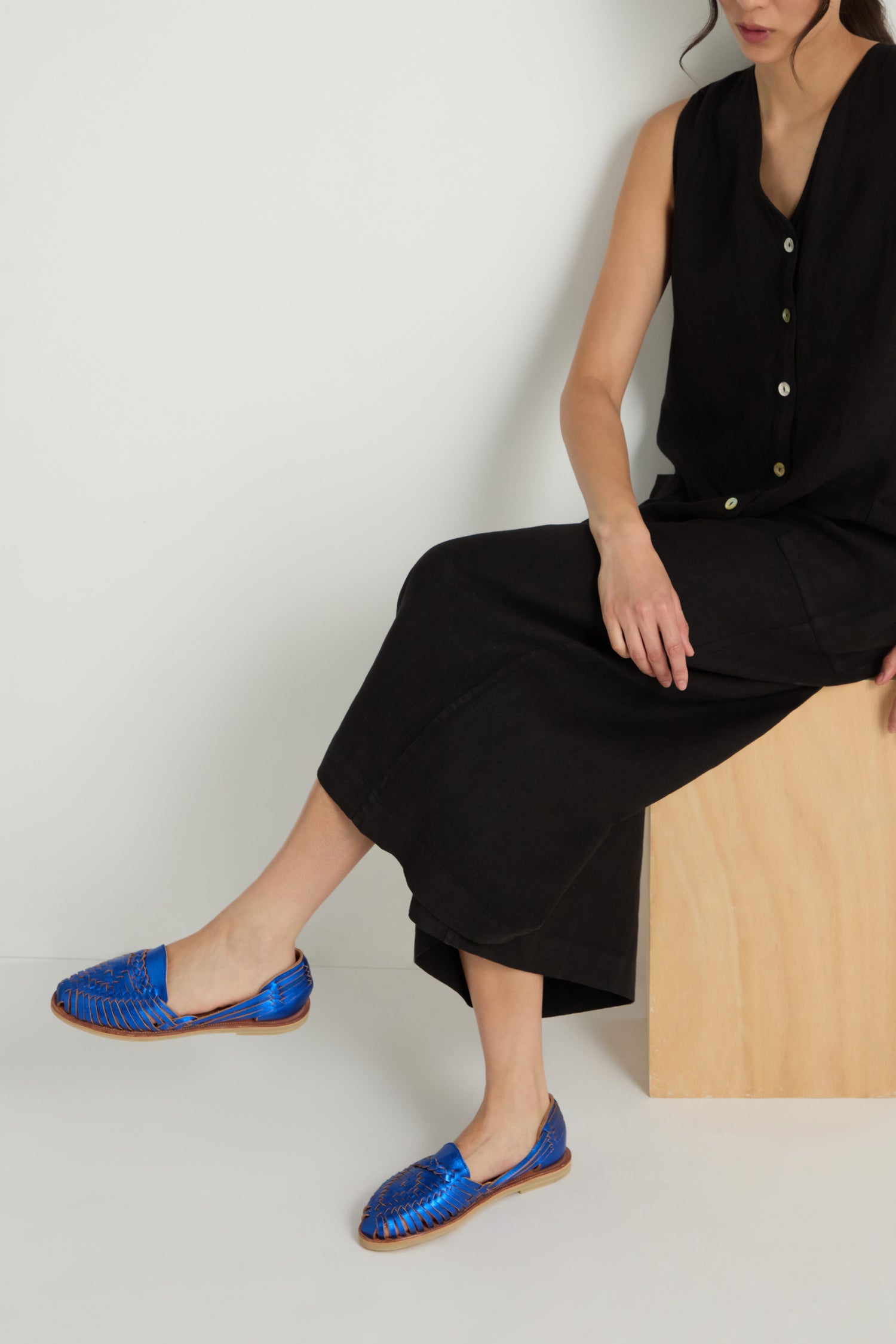 A woman sits on a wooden block, wearing a black sleeveless dress and Metallic Blue Alegre Leather Huarache Sandals, handcrafted in Mexico, against a plain white background.