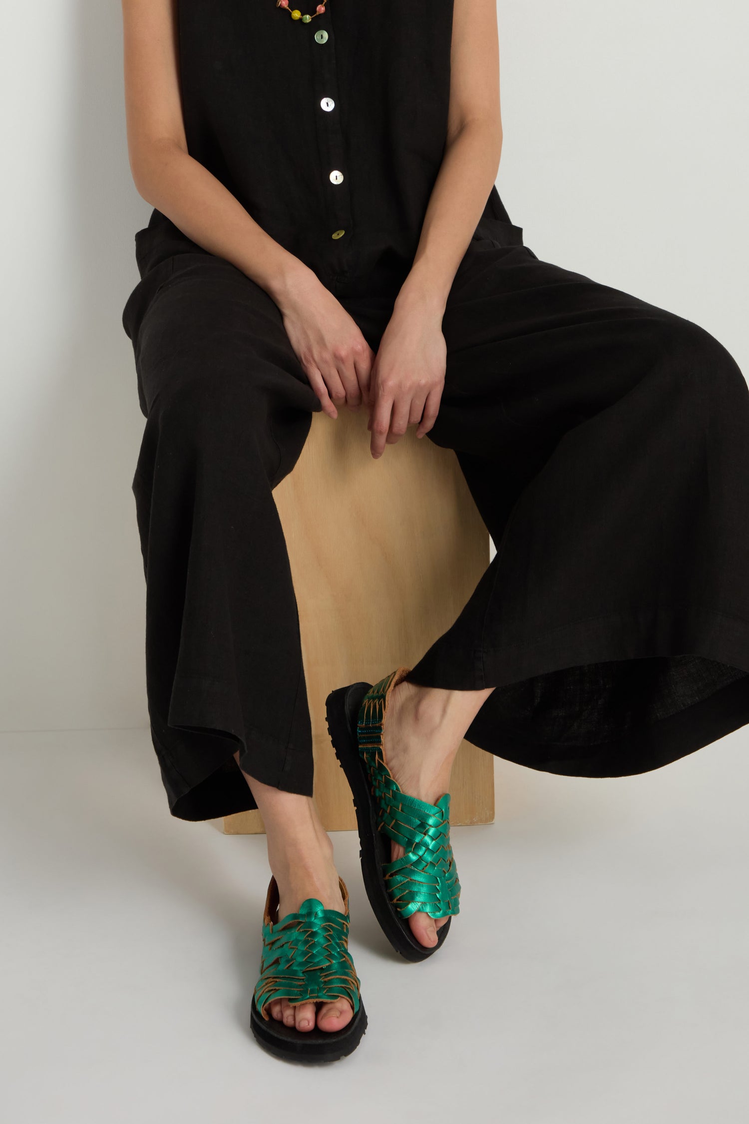 A person sitting on a wooden block wears a black sleeveless outfit and Frida Metallic Green Leather Sandals, handcrafted in Mexico.