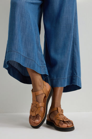 A person in wide-leg denim and Tulum Leather Buckle Sandals stands against a white background, showcasing style with cushioned sole comfort.