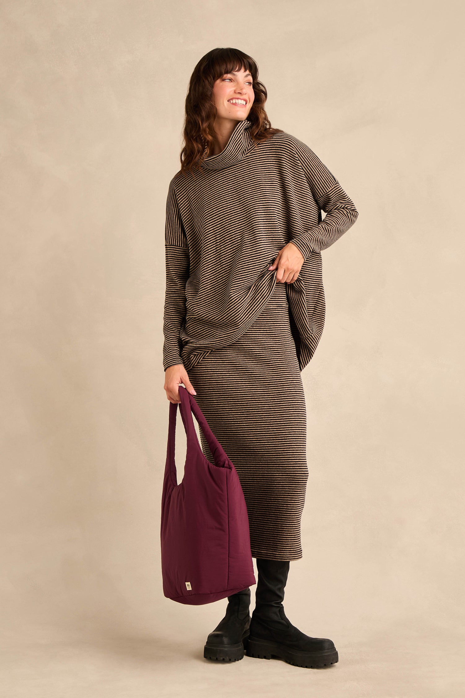 A woman wearing the Roll Neck Narrow Stripe Fleece Tunic paired with black boots and carrying a large purple tote bag stands against a beige background, smiling.