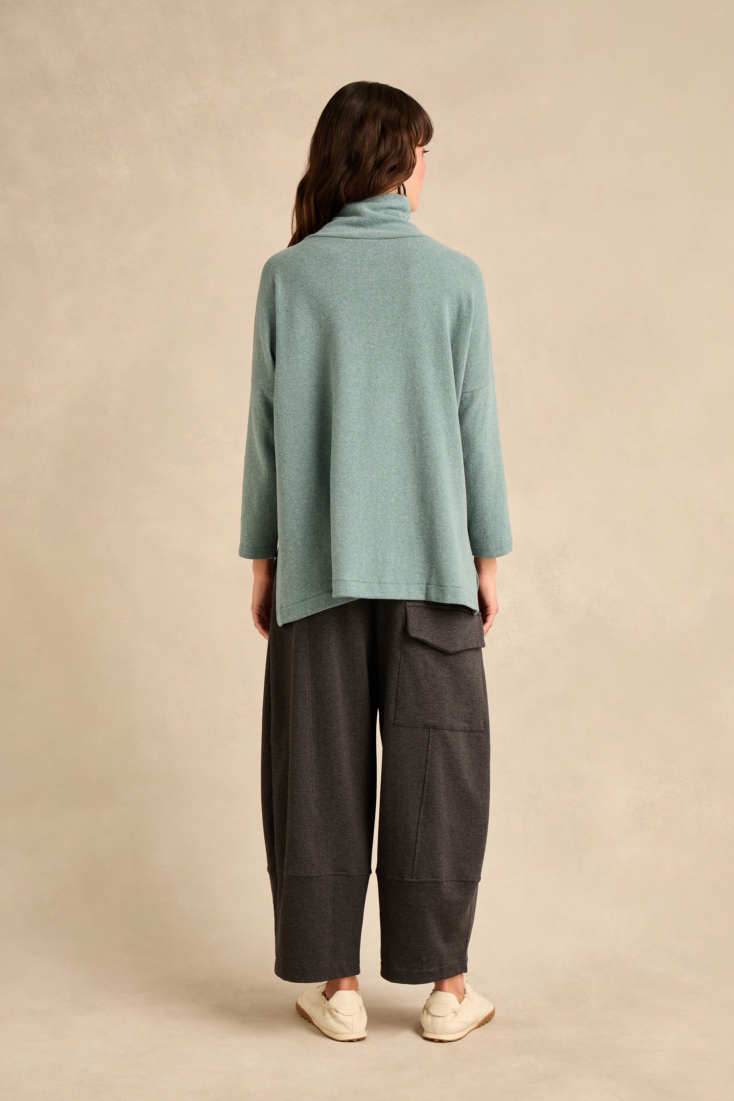 A person stands facing away, wearing the Cowl Neck Soft Jersey Top in a relaxed fit light blue cotton-blend fabric, paired with wide dark gray pants featuring pockets and beige slip-on shoes against a neutral background.