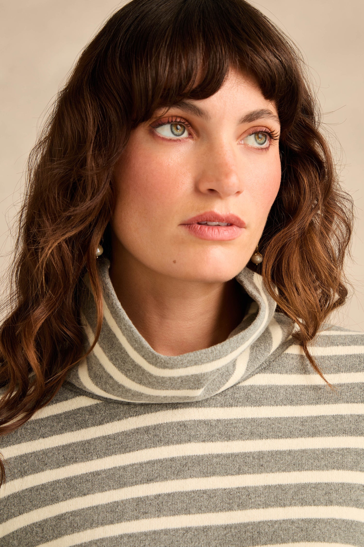 A woman with wavy brown hair is wearing the Roll Neck Silver Wide Stripe Jersey Top, a relaxed-fit style crafted from soft cotton-blend jersey in gray and white wide stripes, as she gazes slightly to the side.
