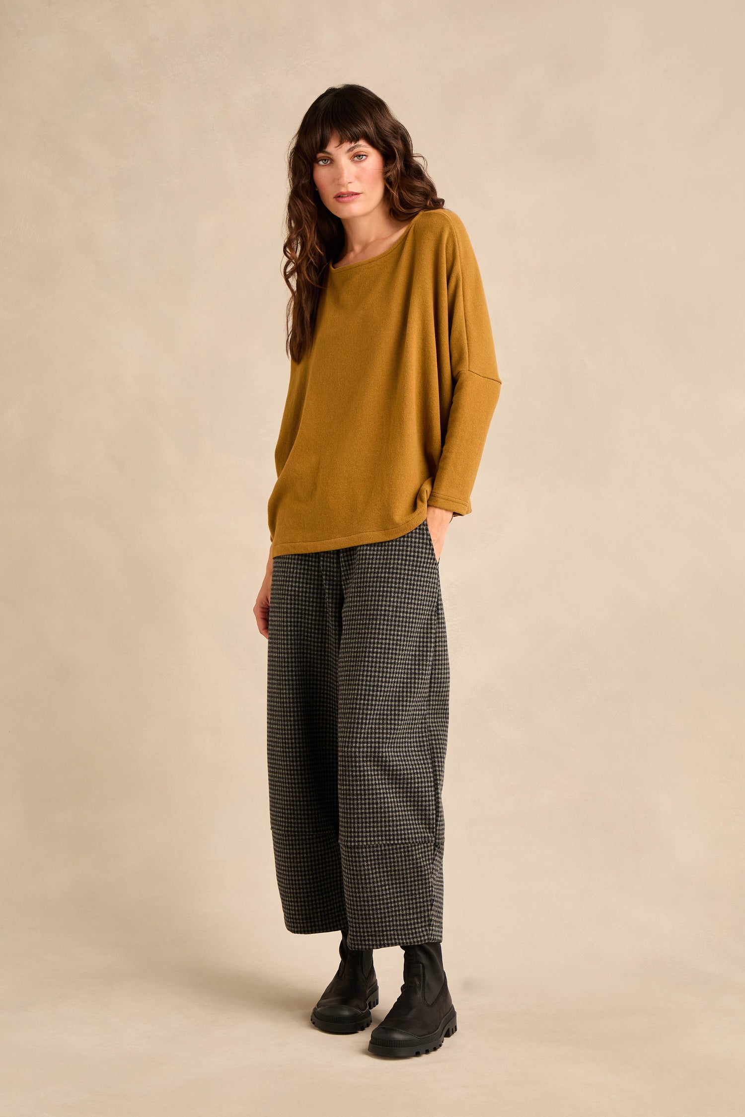 A woman stands against a beige background wearing the Round Neck Soft Jersey Top in mustard, paired with wide-leg checkered pants and black boots.