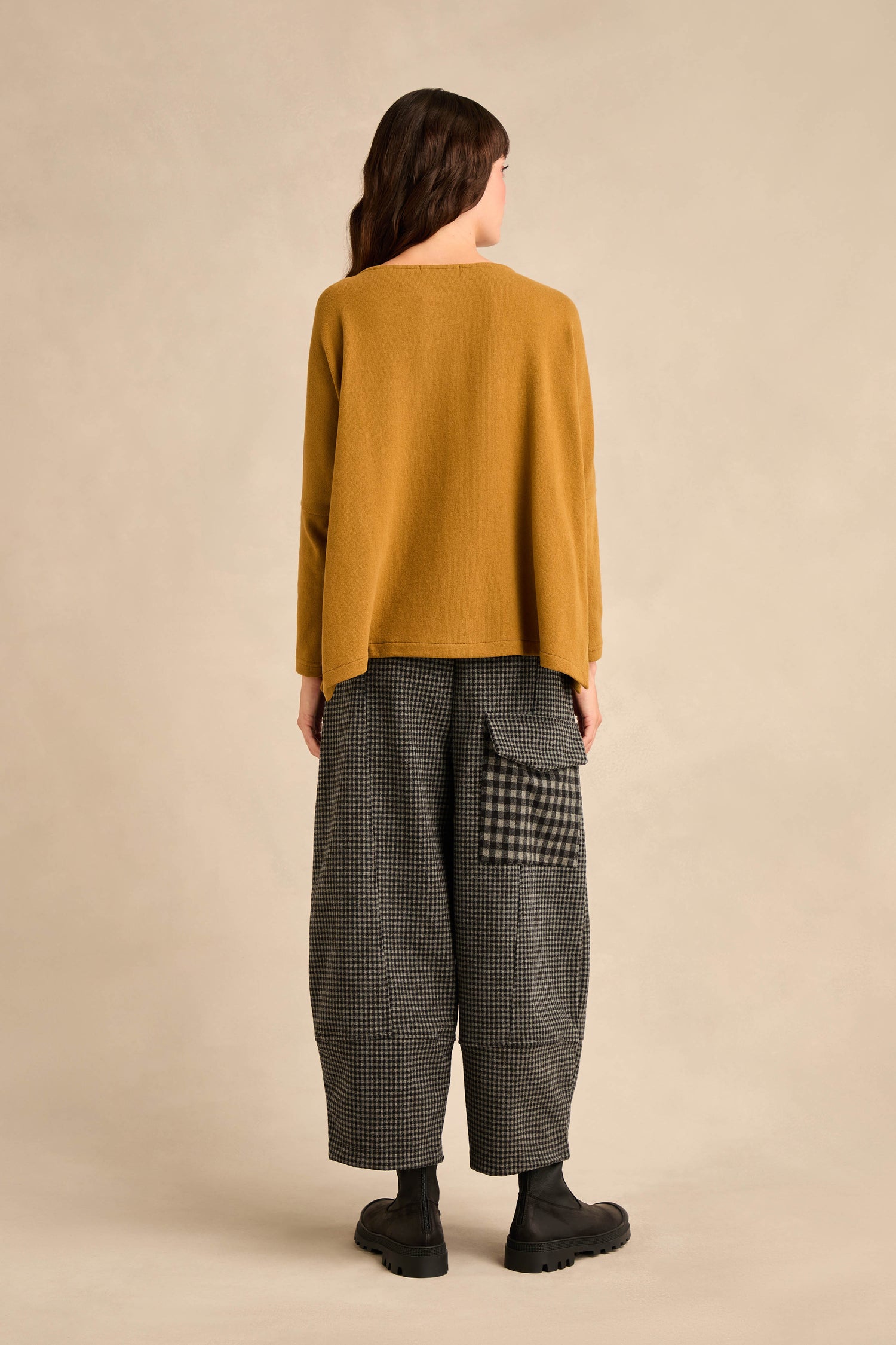 A person with medium-length hair faces away, wearing the Round Neck Soft Jersey Top in mustard—a cotton-blend sweater with a relaxed everyday fit—paired with loose plaid pants featuring a large pocket and black boots, standing against a plain beige background.