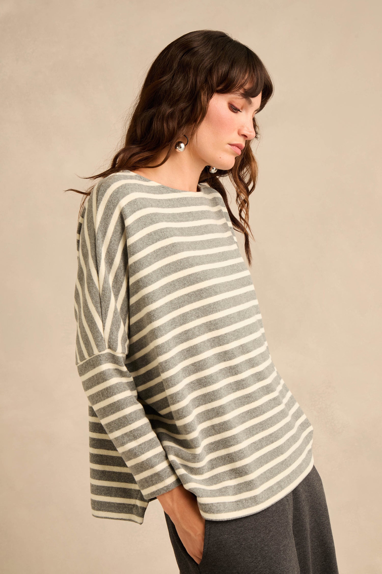 A woman stands against a beige background, looking down, dressed in the Round Neck Wide Stripe Jersey Top—a loose-fitting gray and white striped style featuring a round neckline—paired with dark pants. The cotton-blend jersey fabric adds to the comfortable, casual appeal of her outfit.