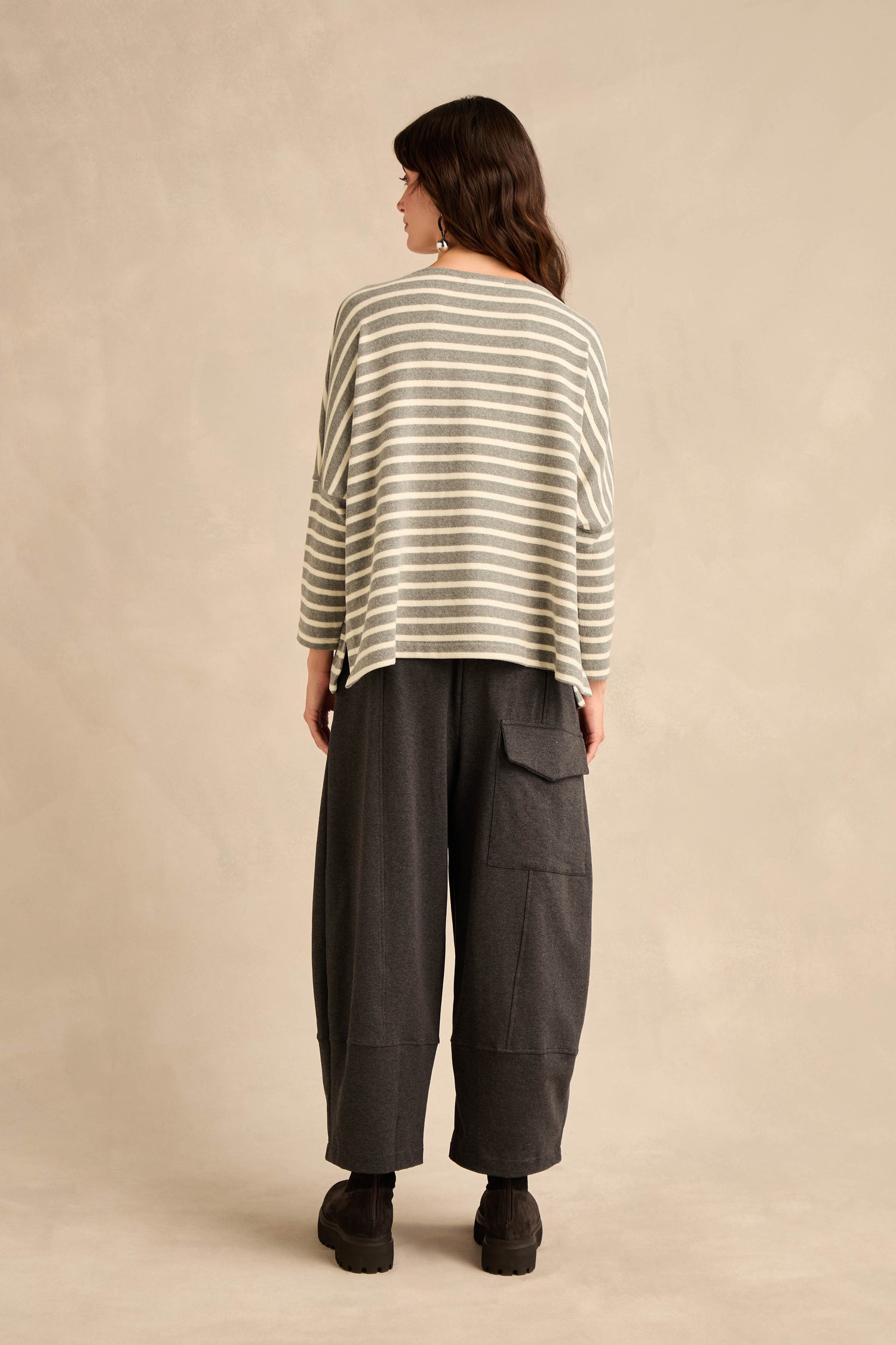A person stands facing away, wearing the Round Neck Wide Stripe Jersey Top crafted from a soft cotton-blend jersey, paired with wide dark pants featuring large pockets and black shoes, against a neutral background.