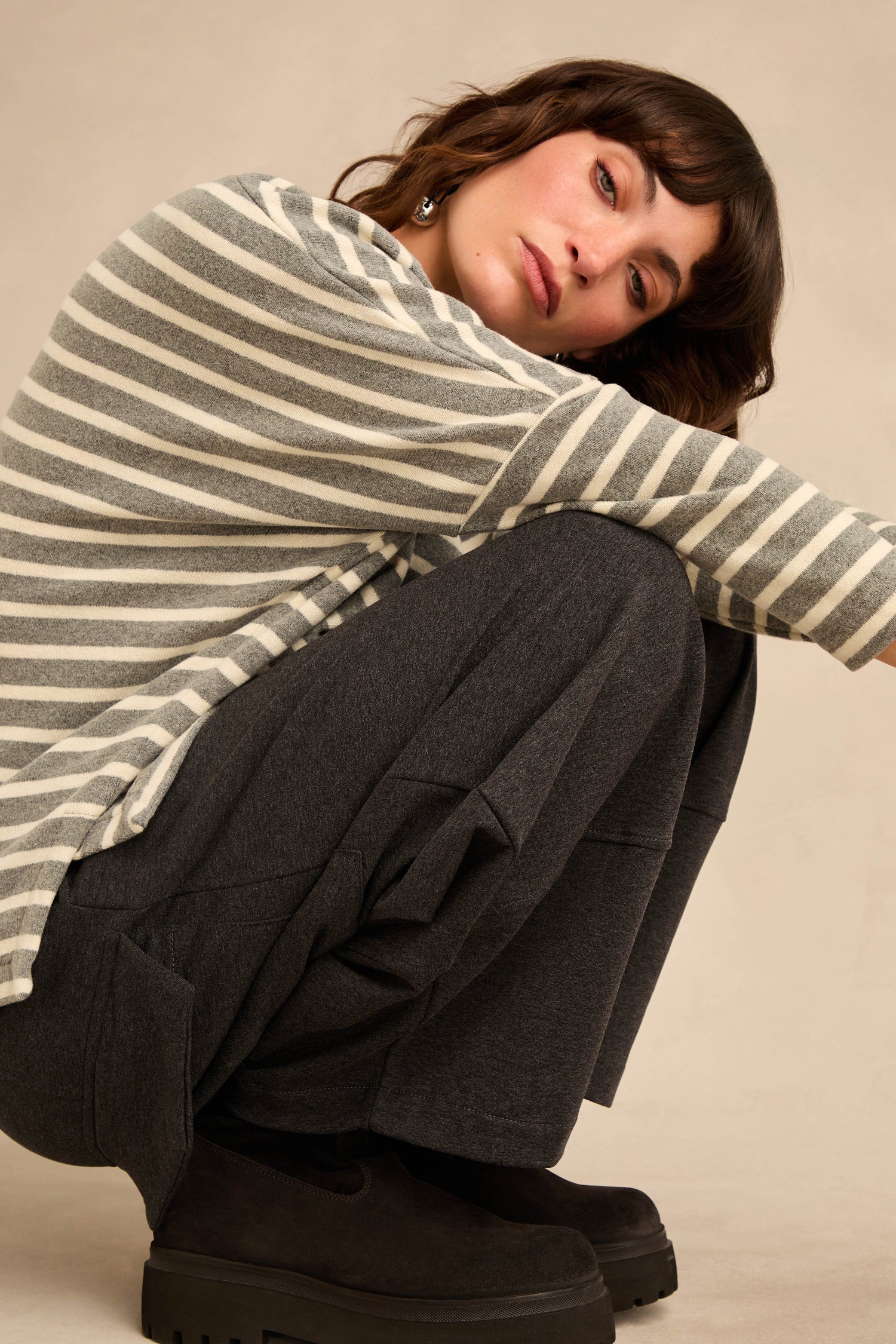 A woman wearing dark Jersey Pocket Trousers and a gray-and-white striped shirt sits on the floor with her knees up and arms wrapped around her legs, looking toward the camera against a plain beige background.