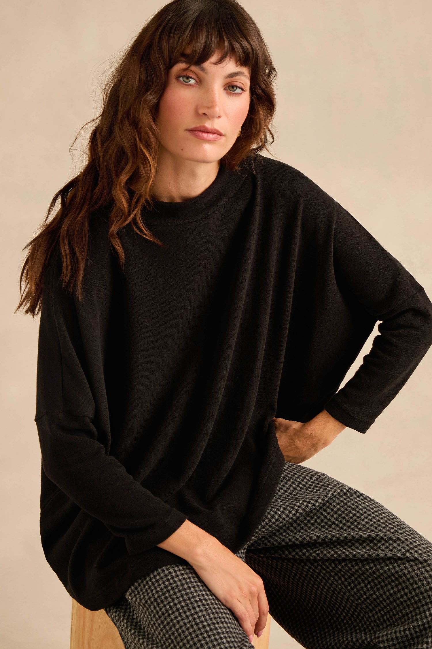 A woman with wavy brown hair sits on a stool against a neutral background, wearing the Black Soft Jersey Boxy Top in a loose, boxy silhouette paired with checkered pants.