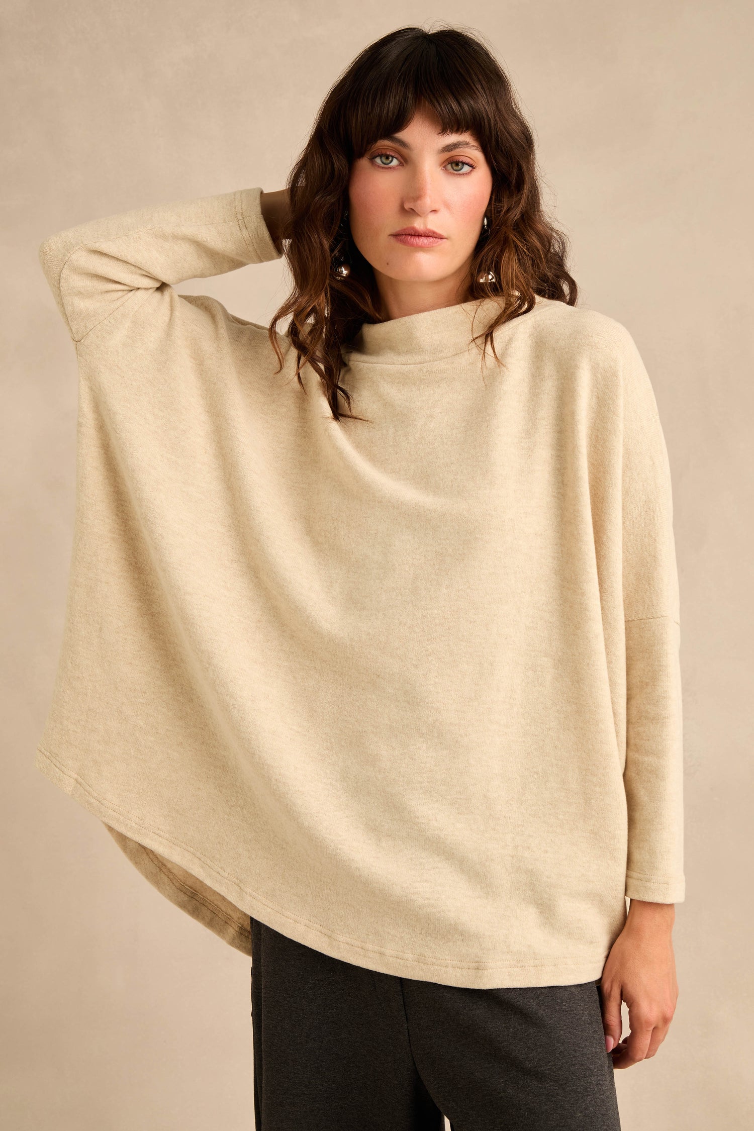 A woman stands against a neutral background wearing the Cream Marl Soft Jersey Boxy Top—a loose, boxy cotton-blend jersey in beige—paired with dark pants, one arm bent and hand touching her head for a relaxed fit look.