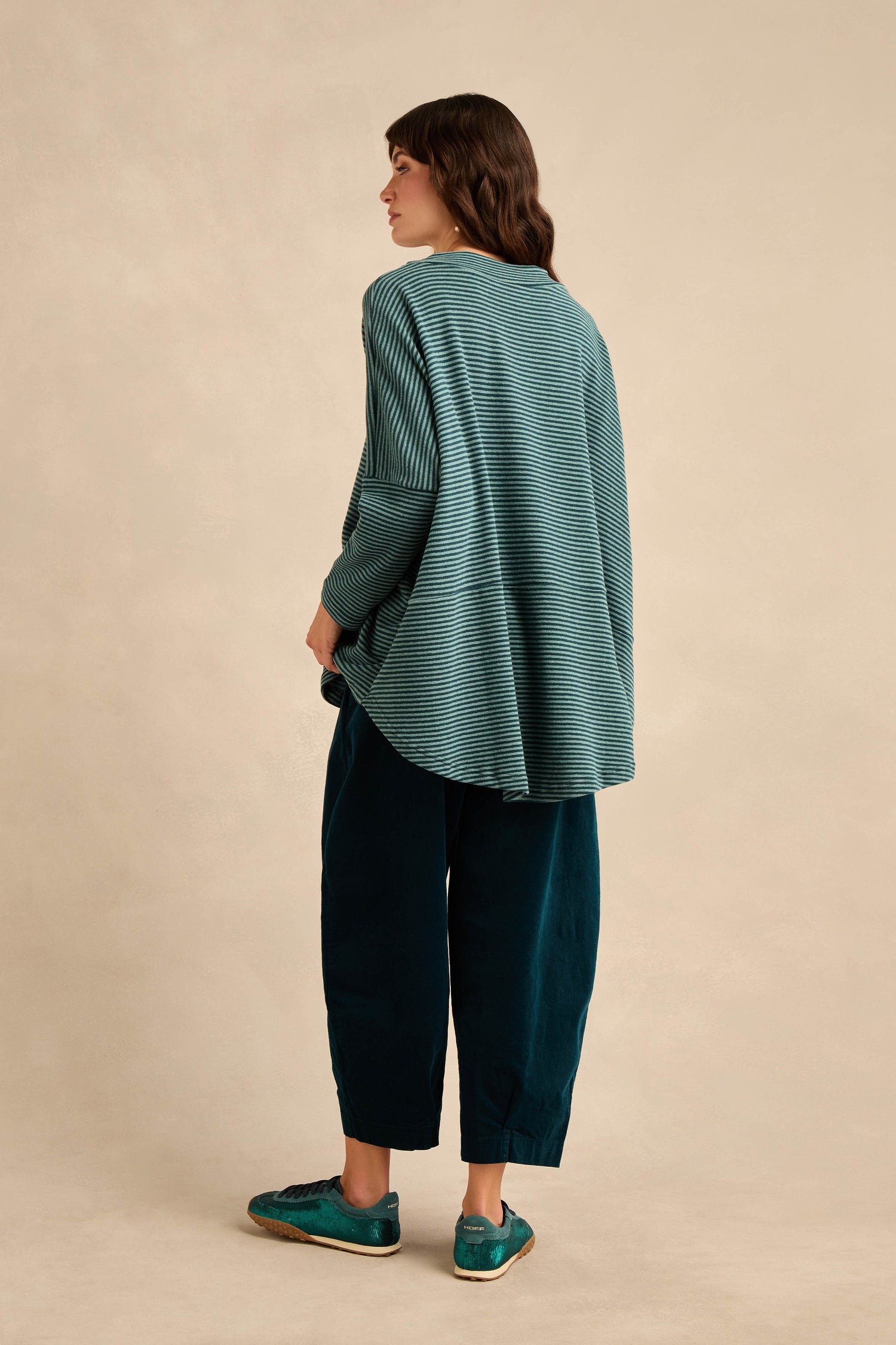 A person stands against a beige background, facing away from the camera, dressed in the Narrow Stripe Soft Jersey Boxy Top—a loose, cotton-blend striped boxy top—paired with wide-leg dark teal pants and teal shoes.