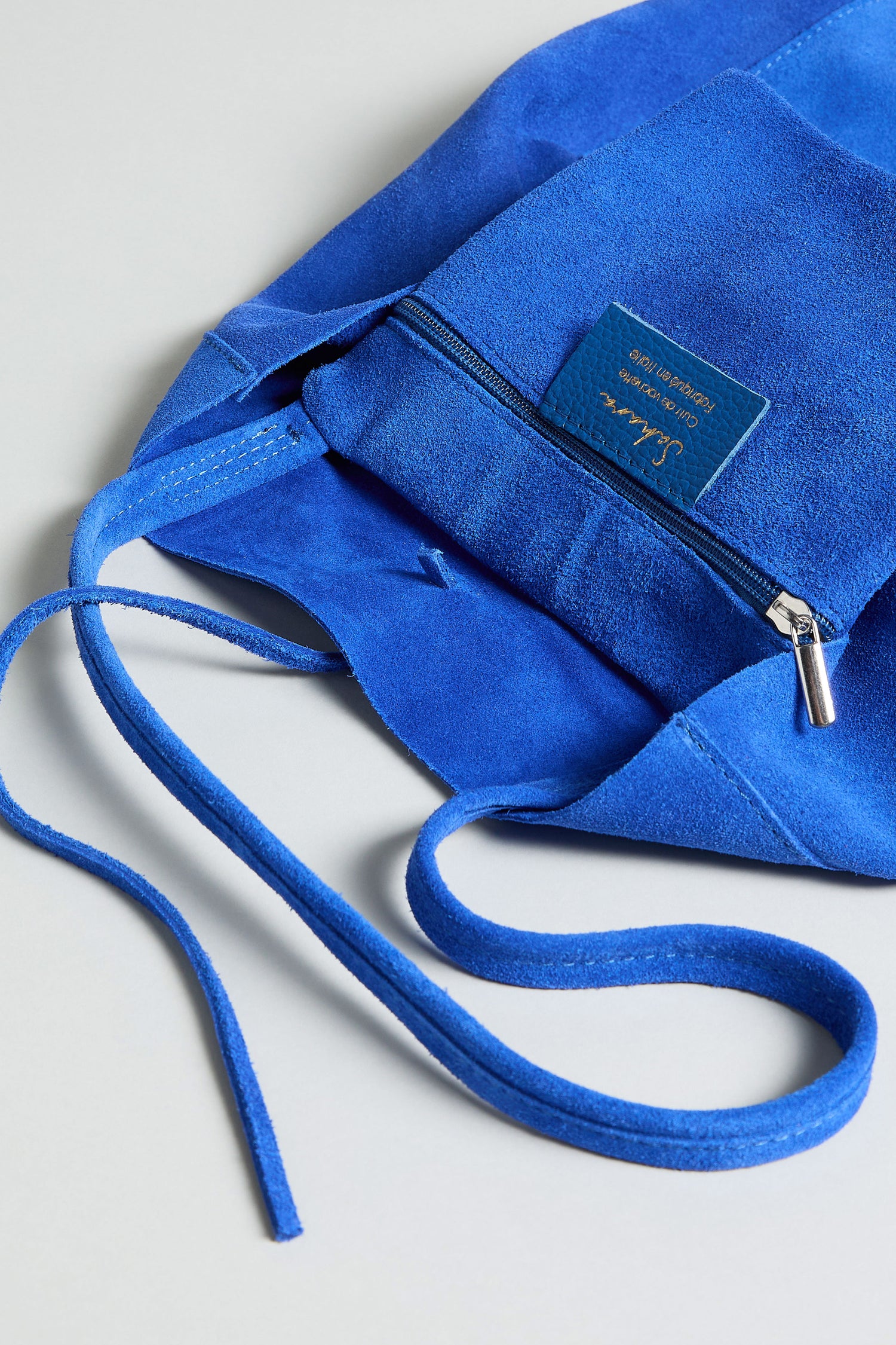 Close-up of the Soft Suede Tote Bag in royal blue, featuring a zippered pocket, visible brand label, and matching suede drawstrings on a light gray surface—an elegant minimalist tote for effortless style.