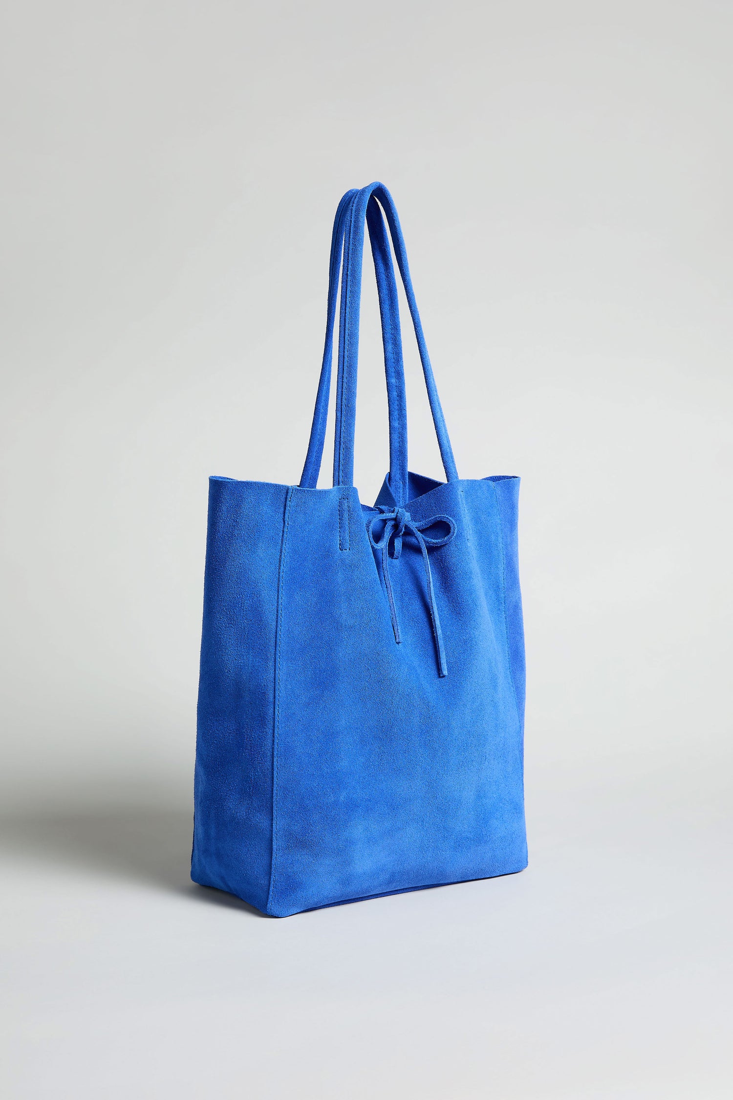 The Soft Suede Tote Bag, in blue with long handles and a front tie detail, is displayed against a plain light background.