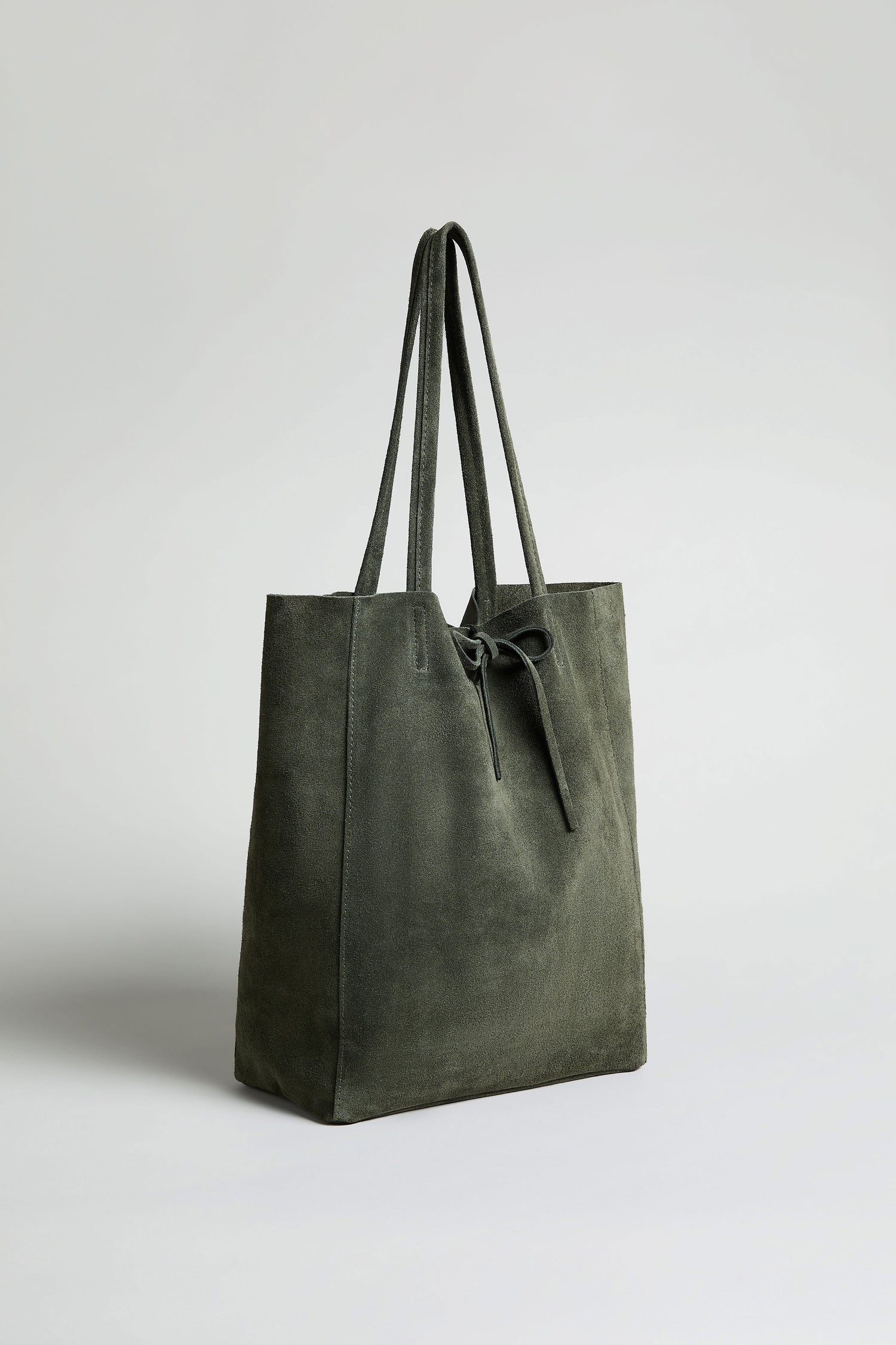 The Soft Suede Tote Bag is a dark green luxury tote in suede with two long handles and a front bow detail, shown against a plain light background.