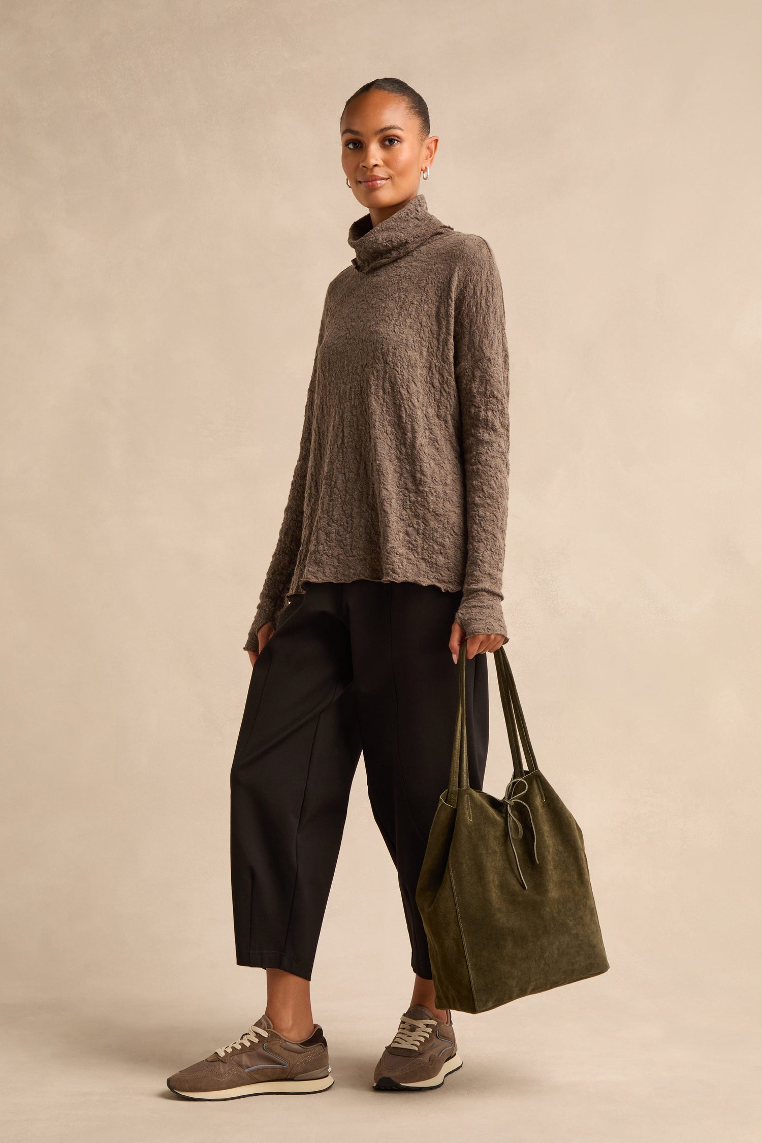 A woman stands against a beige background, carrying the Soft Suede Khaki Tote Bag while wearing a textured brown turtleneck, black pants, and taupe sneakers.